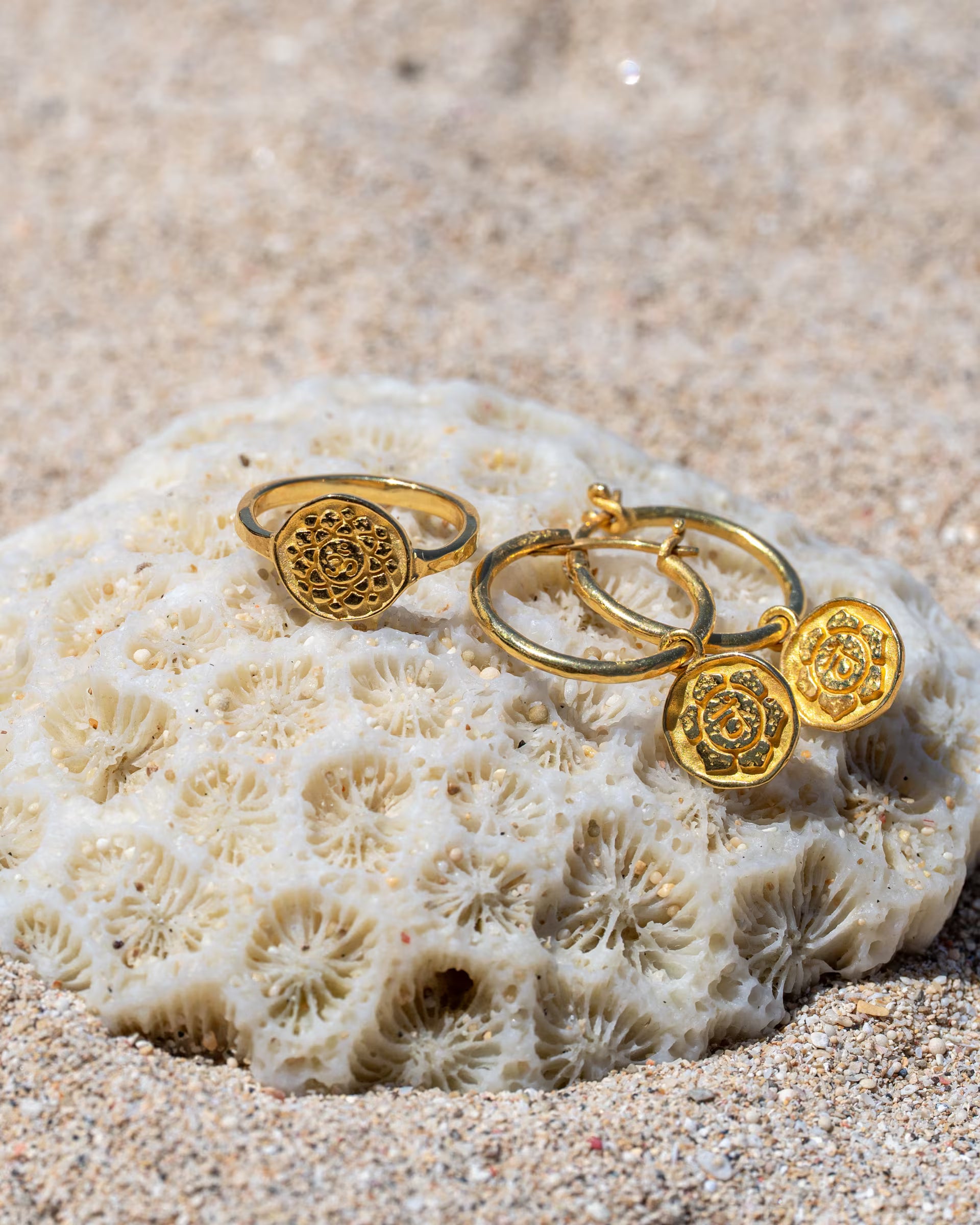 Produktfoto im maritimen Beach-Look von auf Bali handgearbeitetem goldenem Schmuck aus der Chakra-Kollektion – bestehend aus dem CHAKRA RING mit rundem, geprägtem Motiv und den passenden CHAKRA OHRRINGEN als Creolen mit runden Motiv-Anhängern – aus 18k vergoldetem 925er Silber von der Schmuckmarke ILUH, elegant platziert auf einer weißen Koralle im hellen Strandsand.