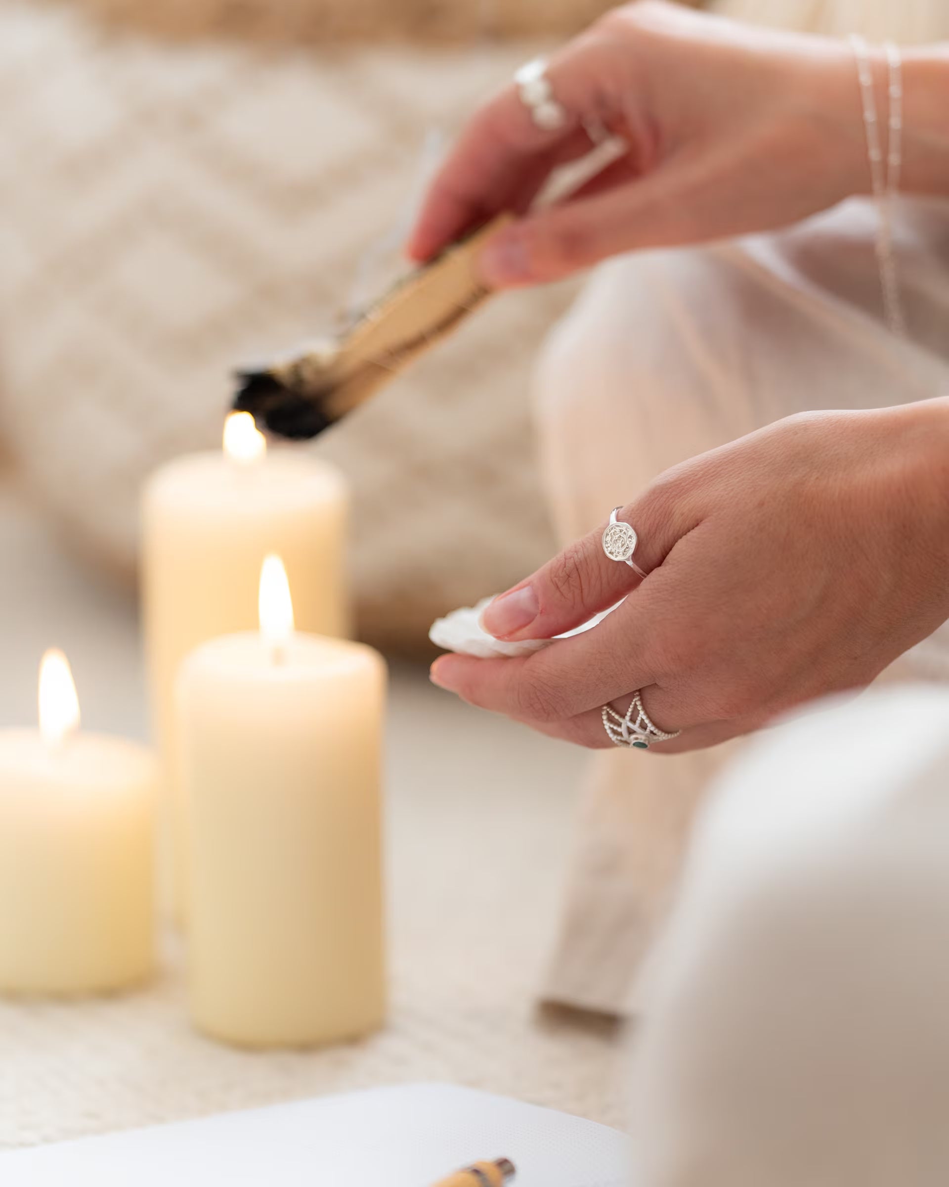 CHAKRA RING SILBER, silberner Schmuck von ILUH, handgefertigt auf Bali – Detailreiche Nahaufnahme im spirituellen Boho-Look von den Händen einer Frau, die ein Palo-Santo-Holzstäbchen an brennenden Kerzen anzündet und dabei eine Kombination aus dem runden CHAKRA RING und dem offenen INTUITION RING mit kleinem Smaragd-Edelstein aus 925er Silber präsentiert.