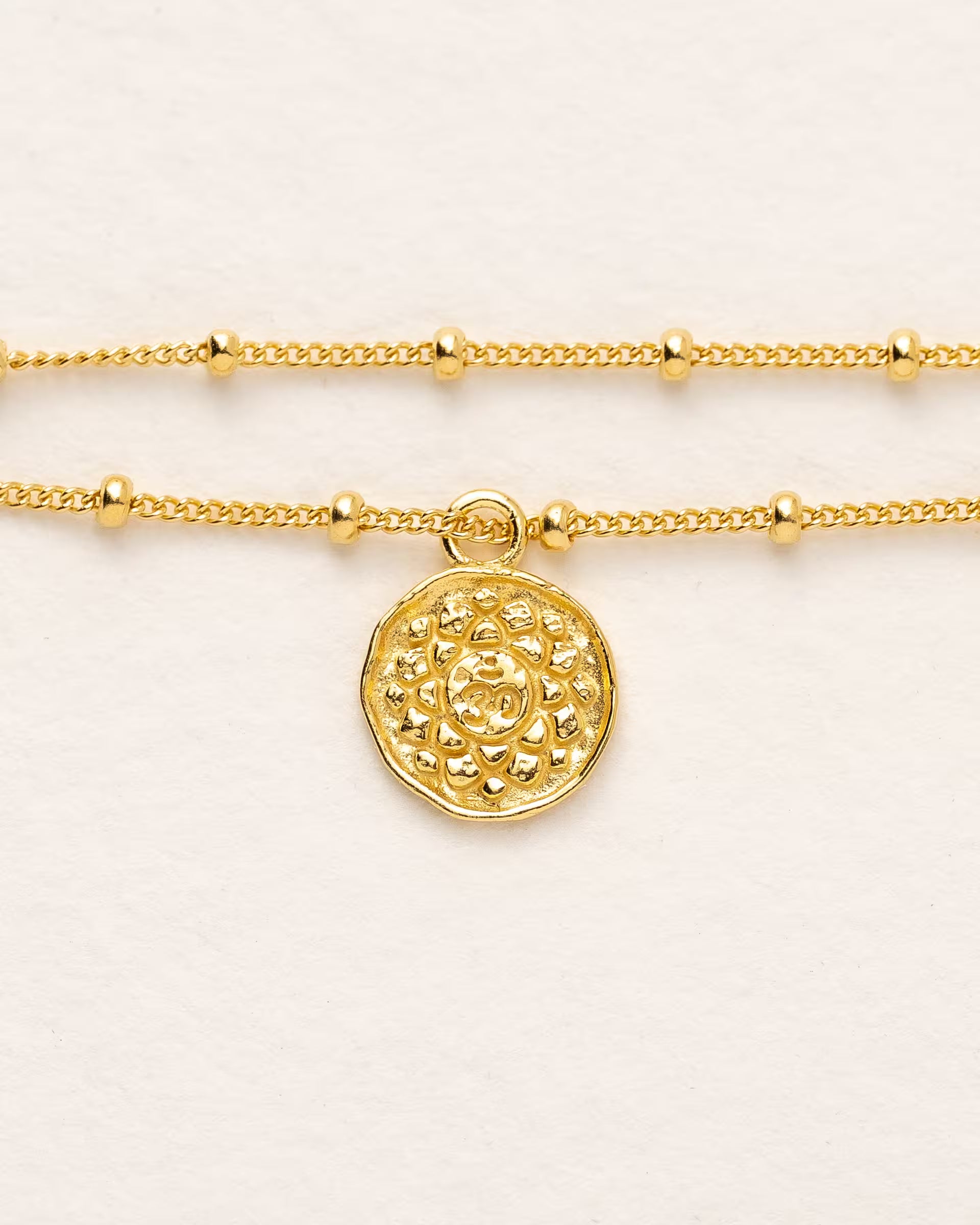 #Motiv_Sahasrara CHAKRA SAHASRARA ARMBAND GOLD, goldenes Armband von ILUH, handgefertigt auf Bali – Spirituelles Produktfoto im minimalistischen Boho-Look von einem feinen Satellitenarmband aus 18k vergoldetem 925er Silber mit einem runden Münzanhänger, der das originale Sanskrit-Zeichen für das Sahasrara-Kronenchakra, eingebettet in kunstvolle Lotusblütenblätter zur Stärkung des Energiezentrums, zeigt, flach präsentiert auf einem hellen, neutralen Büttenpapier-Untergrund.