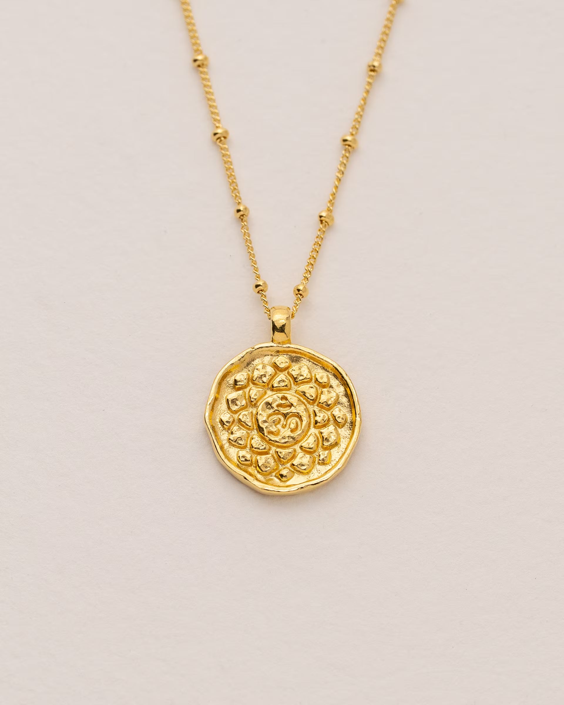 #Motiv_Sahasrara CHAKRA SAHASRARA NECKLACE GOLD, goldene Halskette von ILUH, handgefertigt auf Bali – Spirituelles Produktfoto im minimalistischen Boho-Look von einer feinen Satellitenkette aus 18k vergoldetem 925er Silber mit einem runden Münzanhänger, der das originale Sanskrit-Zeichen für das Sahasrara-Kronenchakra, eingebettet in kunstvolle Lotusblütenblätter zur Stärkung des Energiezentrums, zeigt, flach präsentiert auf einem hellen, neutralen Büttenpapier-Untergrund.