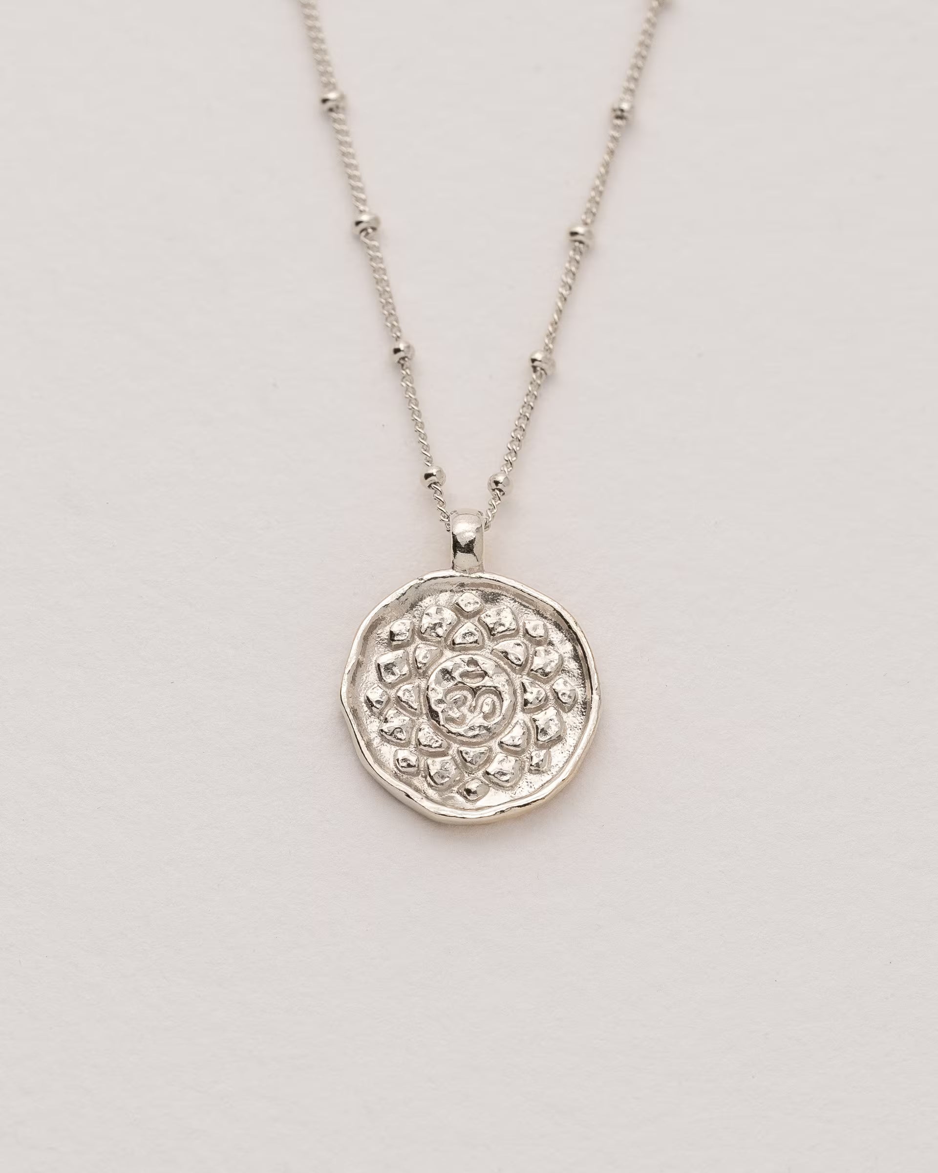 #Motiv_Sahasrara CHAKRA SAHASRARA NECKLACE SILBER, silberne Halskette von ILUH, handgefertigt auf Bali – Spirituelles Produktfoto im minimalistischen Boho-Look von einer feinen Satellitenkette aus 925er Silber mit einem runden Münzanhänger, der das detailliert geprägte Sahasrara-Kronenchakra-Symbol zeigt, flach präsentiert auf einem hellen, neutralen Büttenpapier Untergrund.