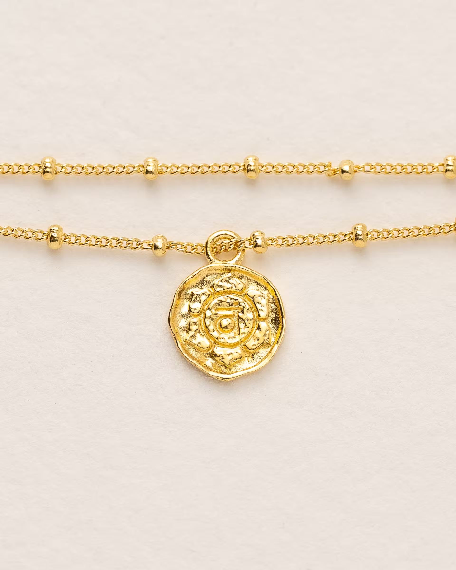 #Motiv_Svadhisthana CHAKRA SVADHISTHANA ARMBAND GOLD, goldenes Armband von ILUH, handgefertigt auf Bali – Spirituelles Produktfoto im minimalistischen Boho-Look von einem feinen, doppelten Satellitenarmband mit einem runden Münzanhänger, der das originale Sanskrit-Zeichen für das Svadhisthana-Sakralchakra, eingebettet in kunstvolle Lotusblütenblätter zur Stärkung des Energiezentrums, zeigt.