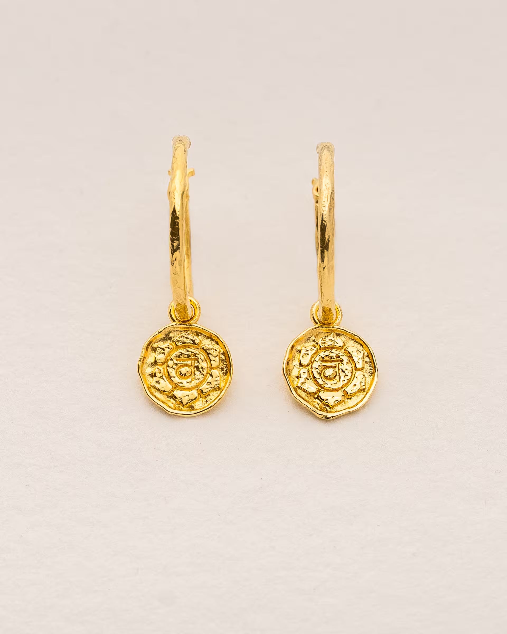 #Motiv_Svadhisthana CHAKRA SVADHISTHANA OHRRINGE GOLD, goldene Ohrringe von ILUH, handgefertigt auf Bali – Spirituelles Produktfoto im minimalistischen Boho-Look von einem Paar feiner Creolen mit einem runden Münzanhänger, der das originale Sanskrit-Zeichen für das Svadhisthana-Sakralchakra, eingebettet in kunstvolle Lotusblütenblätter zur Stärkung des Energiezentrums, zeigt.