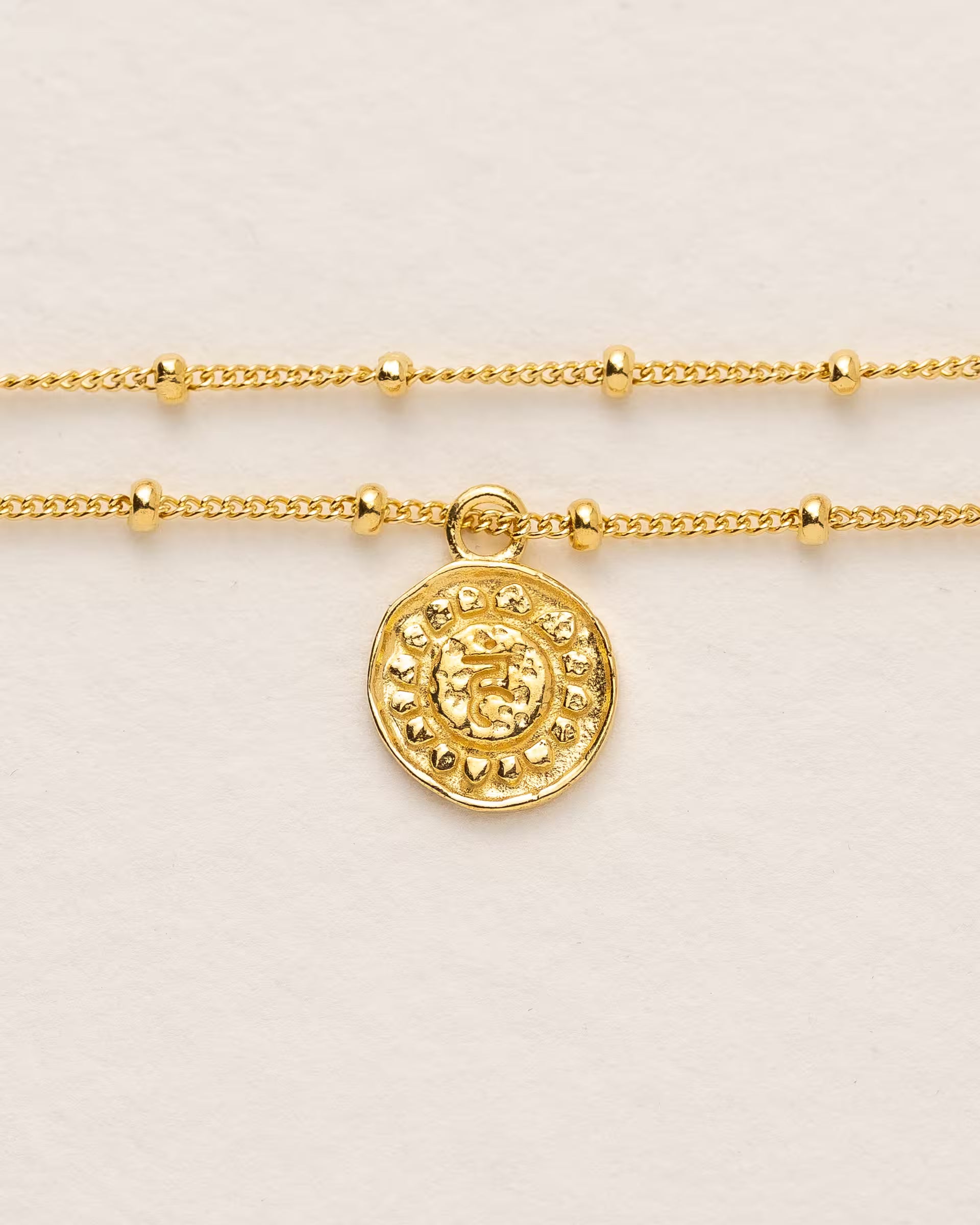 #Motiv_Vishuddha CHAKRA VISHUDDHA ARMBAND GOLD, goldenes Armband von ILUH, handgefertigt auf Bali – Spirituelles Produktfoto im minimalistischen Boho-Look von einem feinen, doppelten Satellitenarmband mit einem runden Münzanhänger, der das originale Sanskrit-Zeichen für das Vishuddha-Halschakra, eingebettet in kunstvolle Lotusblütenblätter zur Stärkung des Energiezentrums, zeigt.