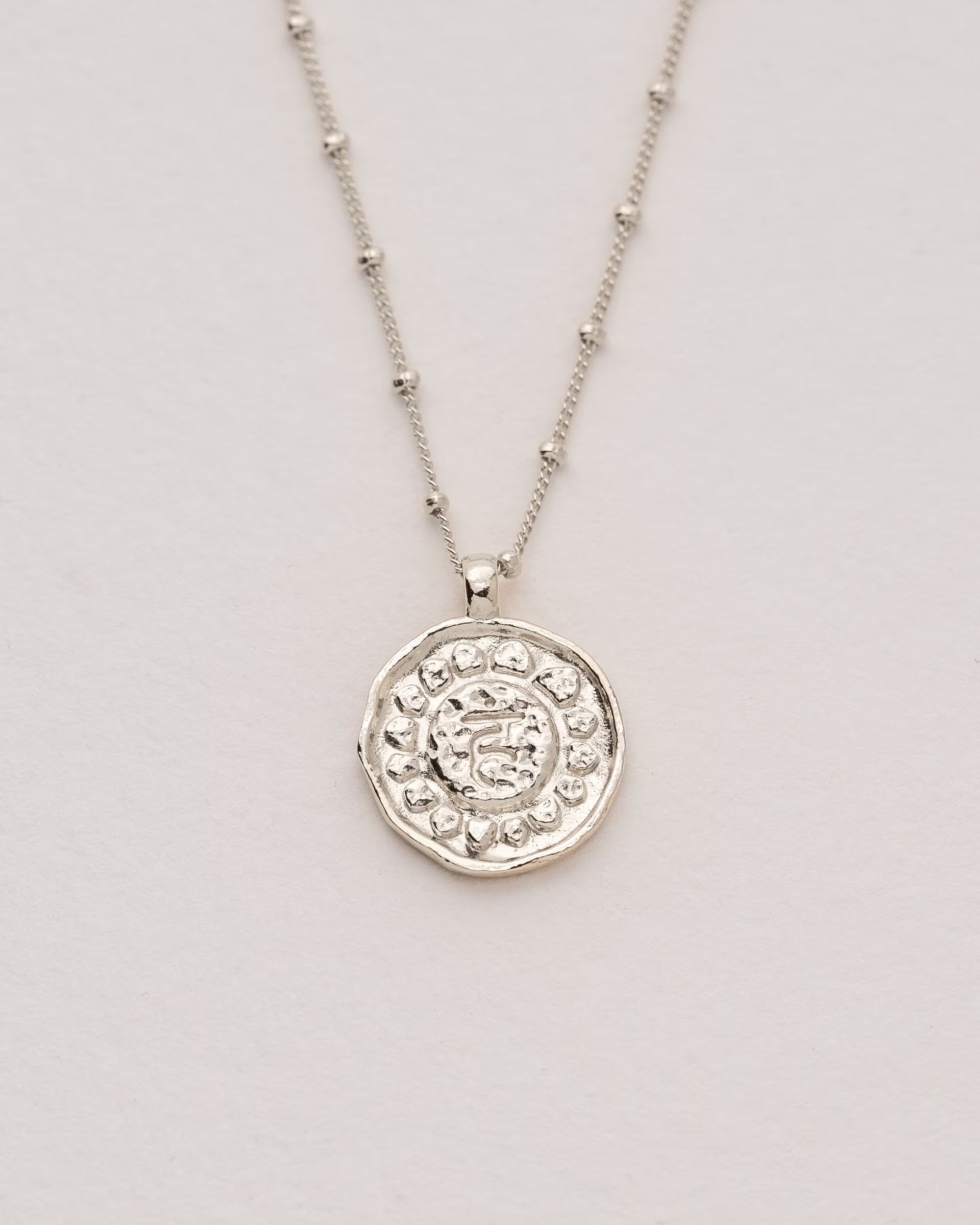 #Motiv_Vishuddha CHAKRA VISHUDDHA NECKLACE SILBER, silberne Halskette von ILUH, handgefertigt auf Bali – Spirituelles Produktfoto im minimalistischen Boho-Look von einer feinen Satellitenkette mit einem runden Münzanhänger, der das originale Sanskrit-Zeichen für das Vishuddha-Halschakra, eingebettet in kunstvolle Lotusblütenblätter zur Stärkung des Energiezentrums, zeigt.