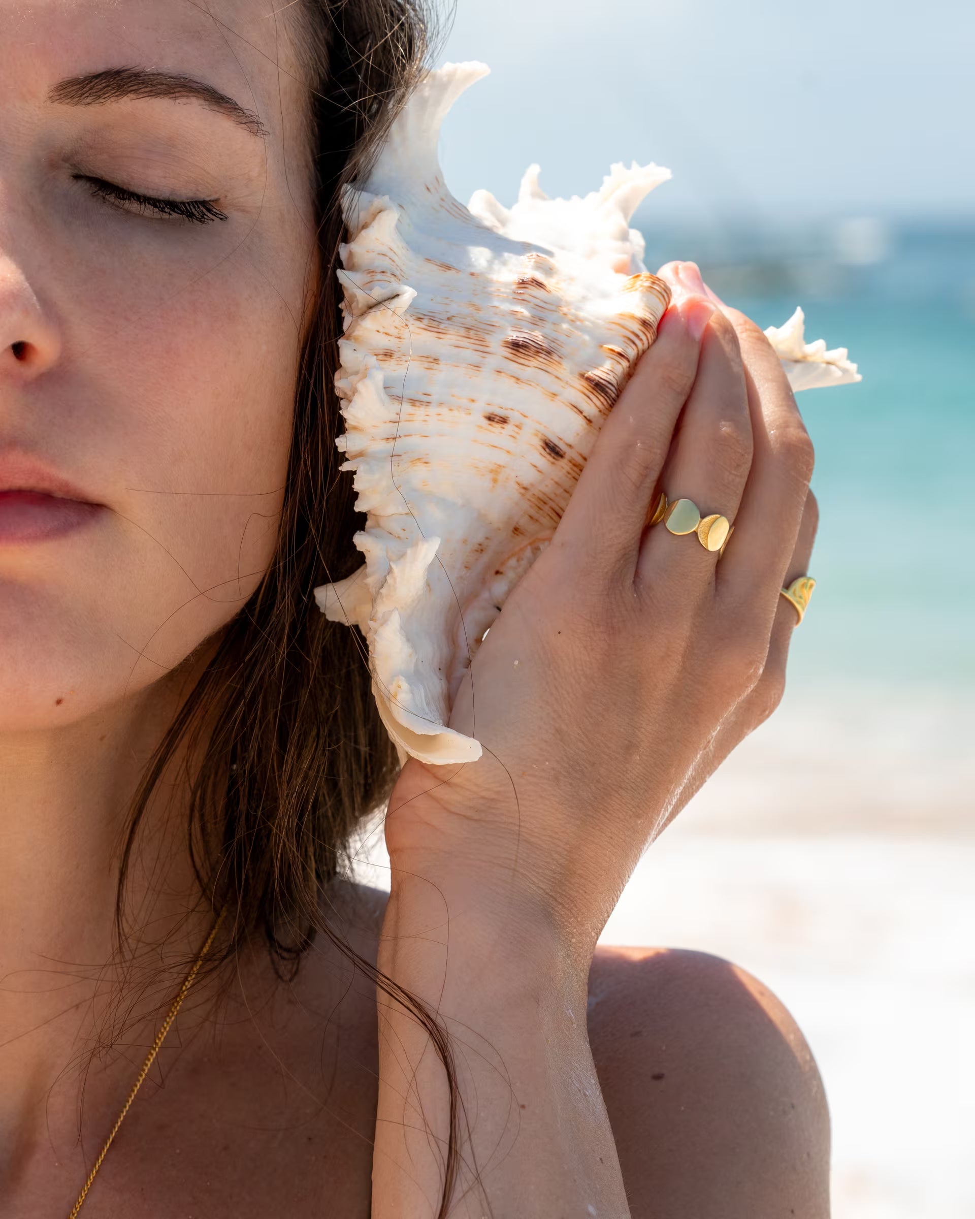 CHANDRA MOONPHASES RING, goldener Schmuck von ILUH, handgefertigt auf Bali – Sinnliche Nahaufnahme am sommerlichen Strand von einer jungen Frau mit geschlossenen Augen, die eine große, strukturierte Tritonshorn-Muschel lauschend an ihr Ohr hält und dabei den breiten, offenen Mondphasen-Ring aus 18k vergoldetem 925er Silber zeigt.