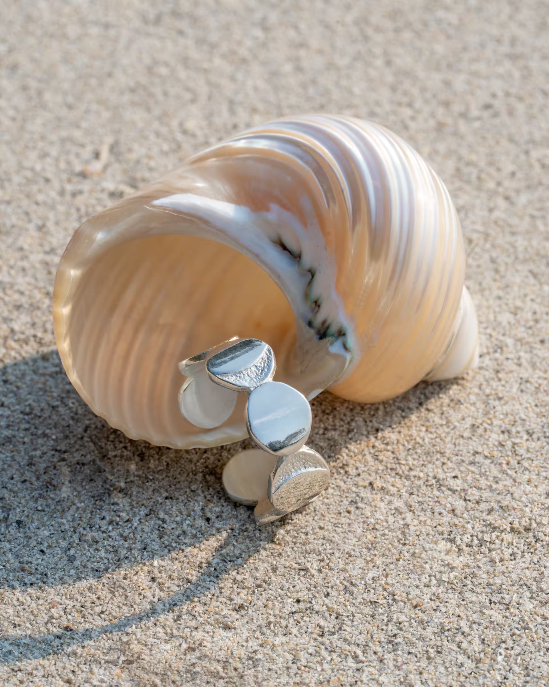 Produktfoto im minimalistischen Beach-Look von dem auf Bali handgefertigten silbernen CHANDRA MOONPHASES RING mit reliefartigen Mondphasen-Motiven aus 925er Silber von der Schmuckmarke ILUH, elegant platziert vor der Öffnung einer großen, gestreiften Strandschnecke im hellen Sand.