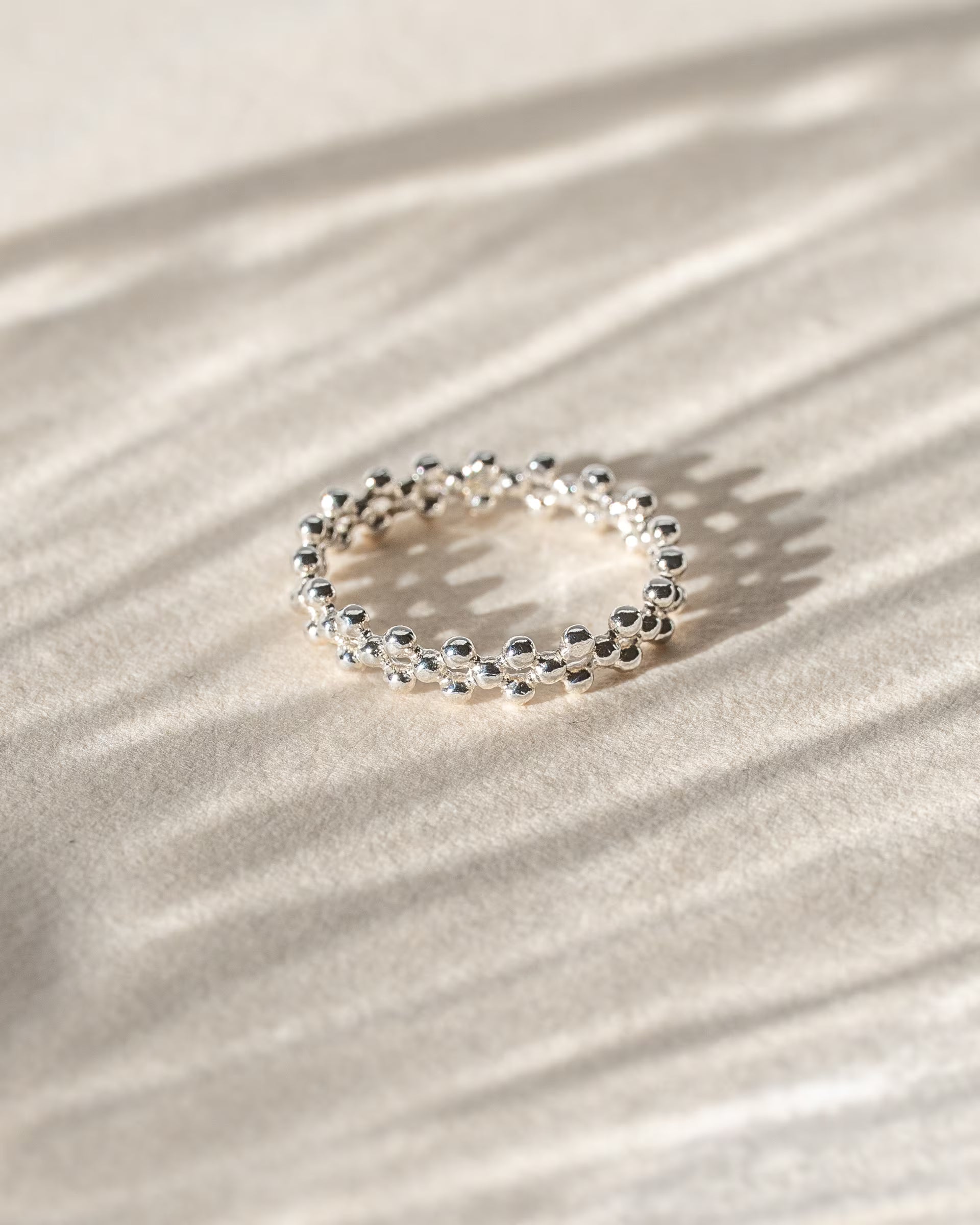 ERIS BEADED RING SILBER, silberner Kugelring von ILUH, handgefertigt auf Bali – Stilvolles Produktfoto im minimalistischen Boho-Look von einem strukturierten Ring aus 925er Silber, der aus zwei Reihen versetzt aneinandergereihte, feine Silberkügelchen besteht, effektvoll präsentiert im lebendigen Streiflicht mit markantem Schattenspiel auf einem hellen, neutralen Büttenpapier Untergrund.