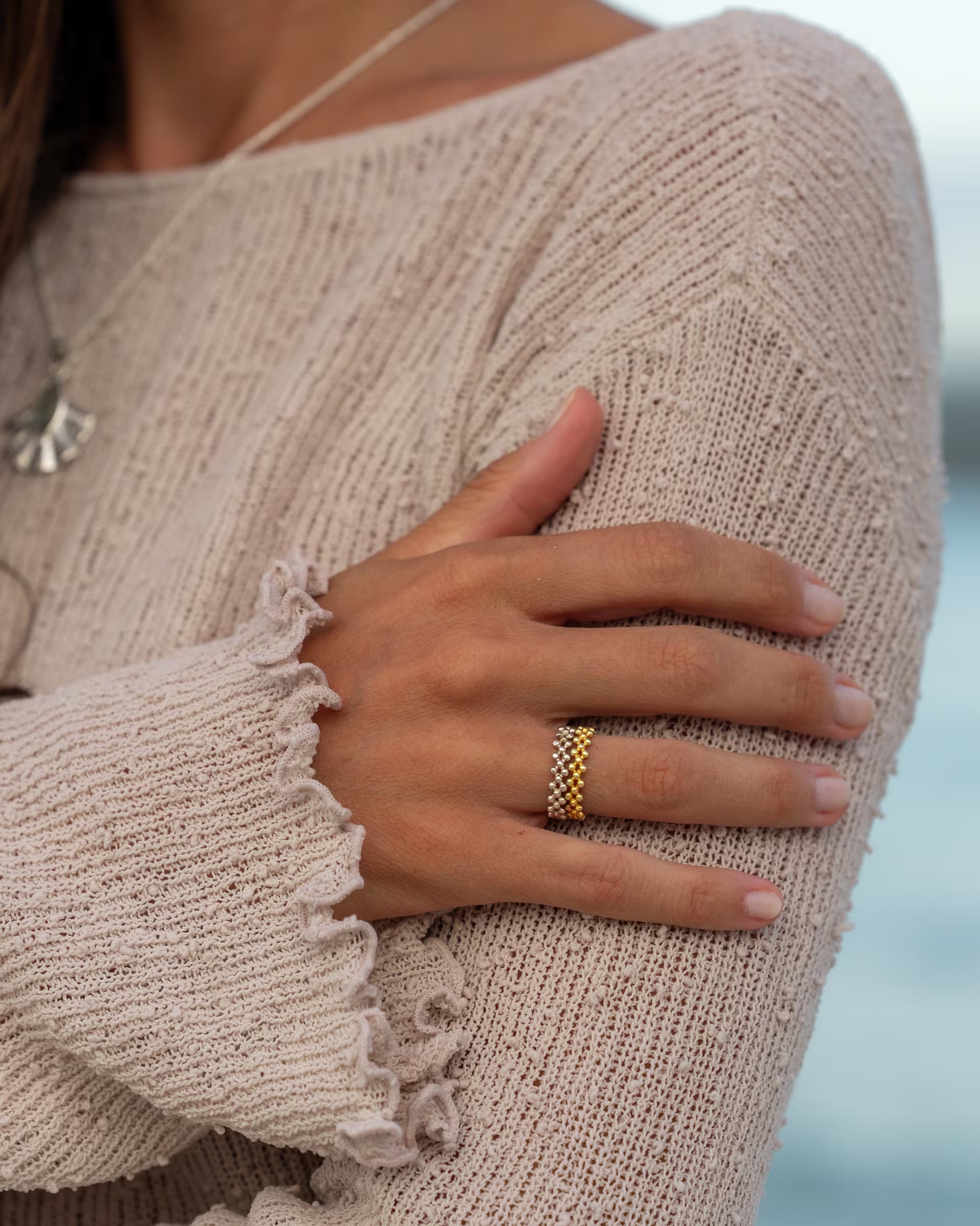 Handgefertigter ERIS Ring im silber-goldenen Stacking-Look aus 925er Silber und 18k Vergoldung von der Schmuckmarke ILUH, von einer jungen Frau getragen am Meer zum beigen Strickpullover mit der edlen ALUN Halskette mit Labradorit-Edelstein im Hintergrund.