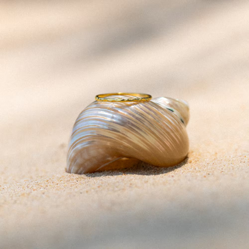 Stimmungsvolle Nahaufnahme eines minimalistischen Rings der Schmuckmarke ILUH, drapiert auf einer glänzenden Muschel im Sand. Das handgefertigte Schmuckstück aus recyceltem 925 Sterling Silber ist mit 18k Echtgold in Deutschland veredelt. Ein perfektes Accessoire für den sommerlichen Beach Vibes Look, das balinesische Handwerkskunst mit zeitloser Eleganz verbindet.