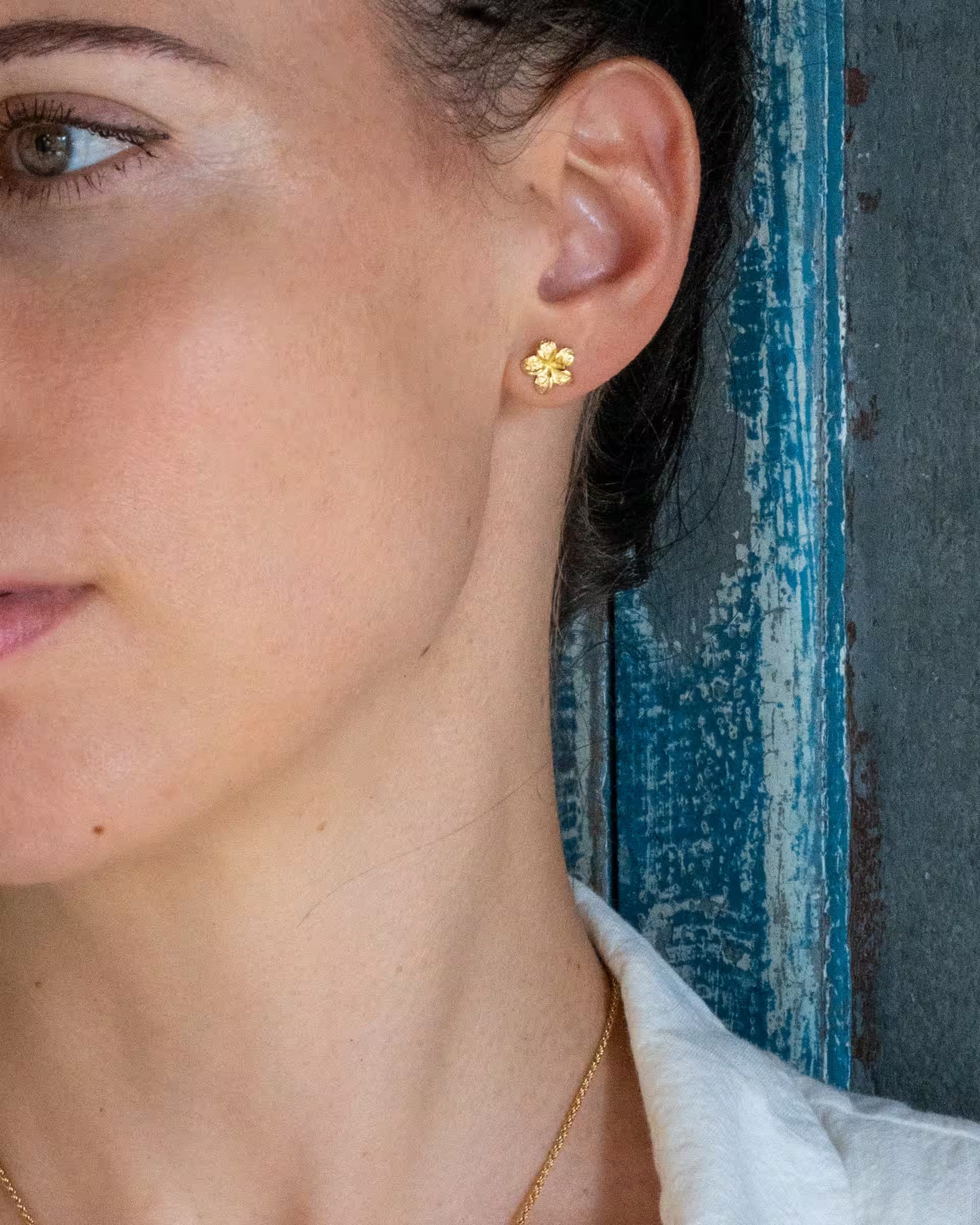 Nahaufnahme im rustikalen Boho-Stil von einer Frau im Profil vor einer blau verwitterten Holzwand, die die auf Bali handgefertigten goldenen FRANGIPANI OHRSTECKER in Form einer filigranen Frangipani-Blüte aus 18k vergoldetem 925er Silber von der Schmuckmarke ILUH trägt.