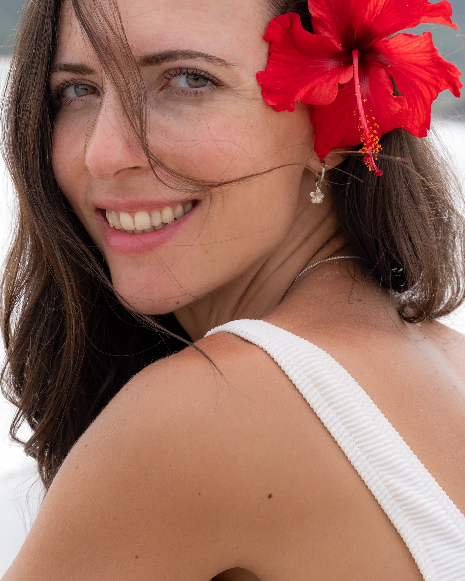 Nahaufnahme im tropischen Beach-Look von einer herzlich lächelnden Frau im weißen Rippstrick-Badeanzug, die über die Schulter blickt und eine leuchtend rote Hibiskusblüte im Haar trägt, kombiniert mit auf Bali handgefertigtem silbernem Schmuck – bestehend aus den kleinen FRANGIPANI OHRRINGEN als Creolen mit zarten Blüten-Anhängern – aus 925er Silber von der Schmuckmarke ILUH.