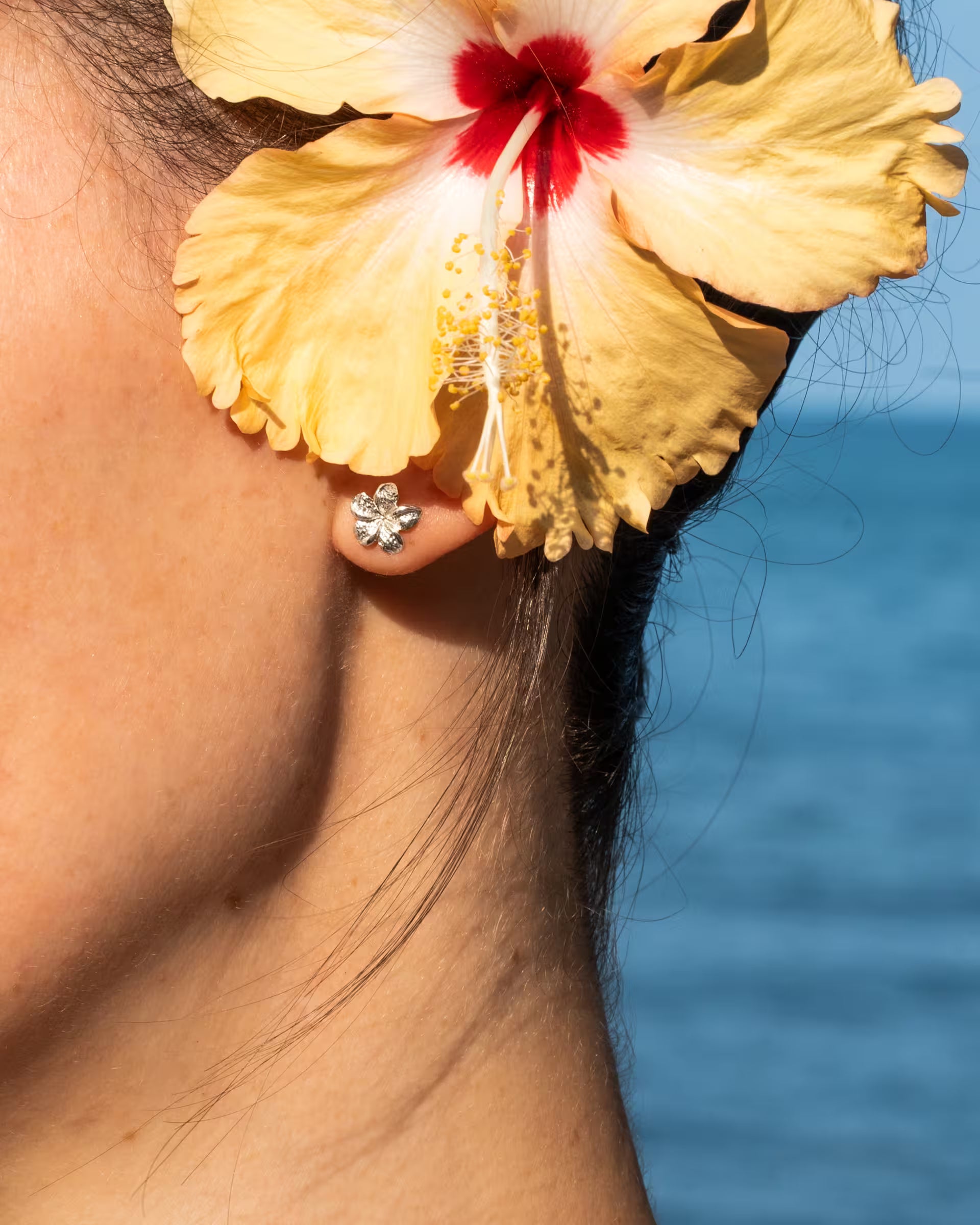 Handgefertigte Frangipani Ohrstecker aus 925er Silber von der Schmuckmarke ILUH, getragen von einer Frau mit einer gelb-roten Hibiskusblüte im Haar vor blauem Meer im Sommer-Look.