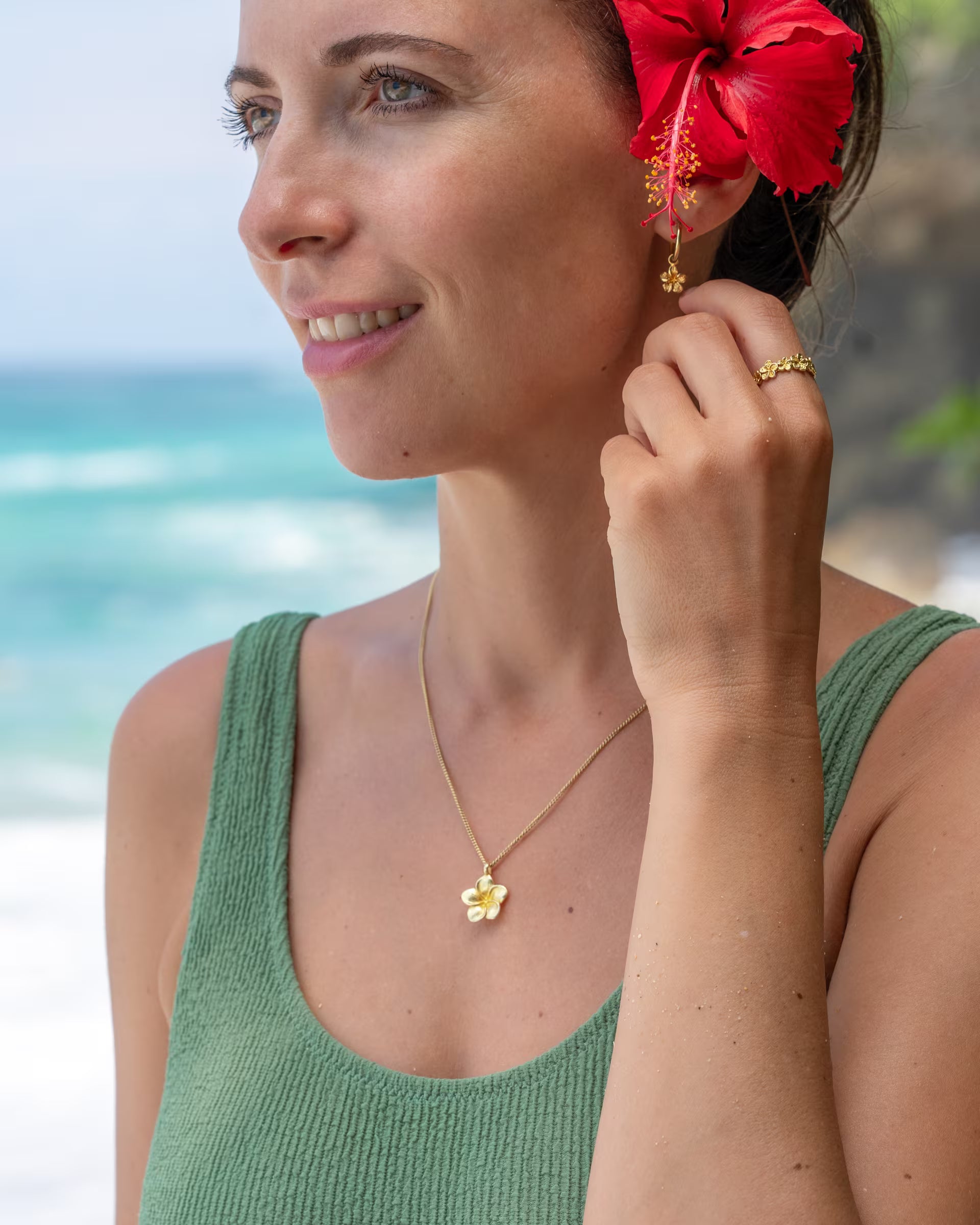 FRANGIPANI FLOWER SCHMUCKSET, goldene Halskette, Ohrringe und Ring von ILUH, handgefertigt auf Bali – Tropische Nahaufnahme von einer lächelnden Frau im salbeigrünen Badeanzug am Meer mit einer roten Hibiskusblüte im Haar, die den detailreichen Frangipani-Blütenanhänger, die dazu passenden kleinen Creolen sowie den strukturierten Frangipani RING aus 18k vergoldetem 925er Silber präsentiert.