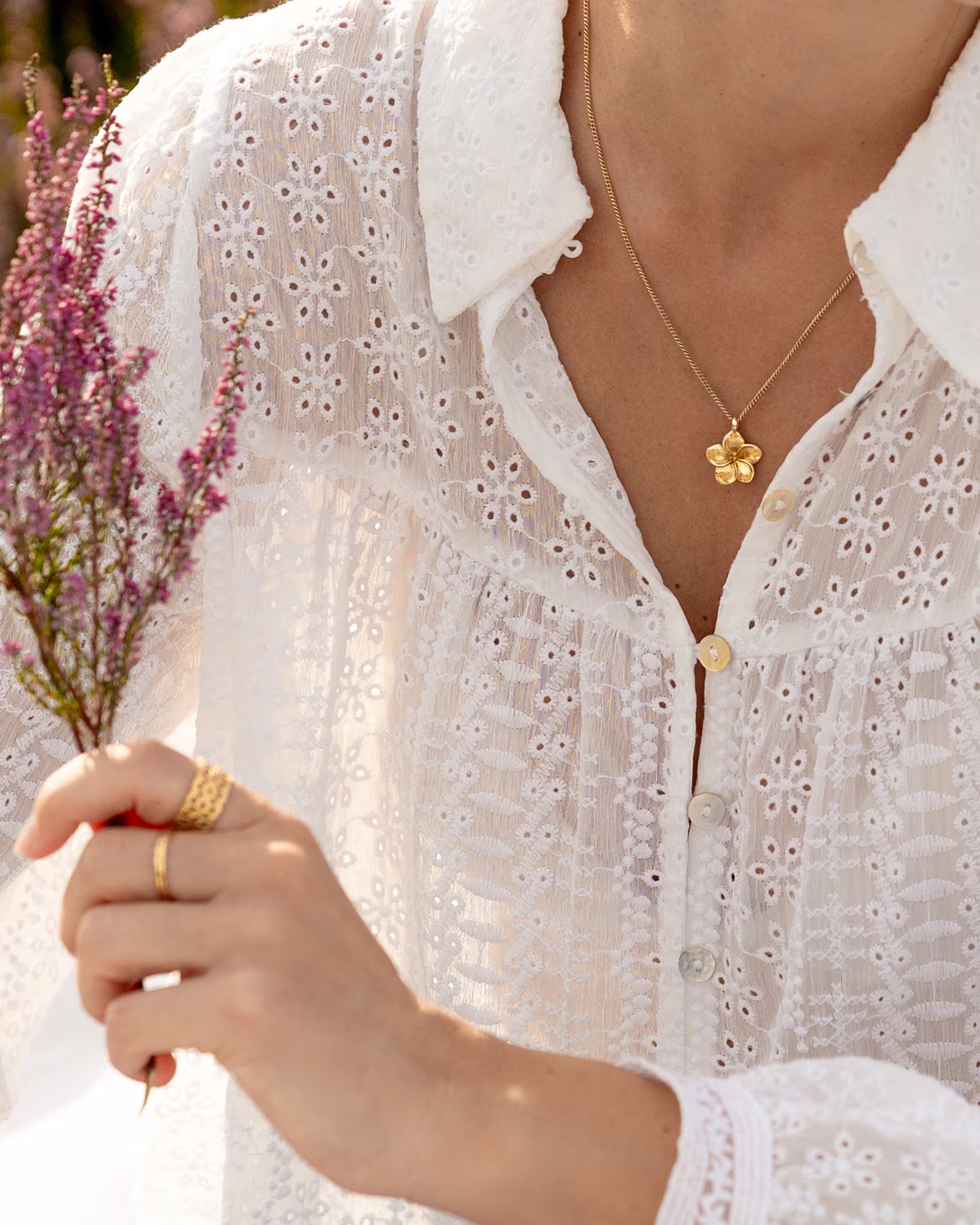 FRANGIPANI KETTE, goldener Schmuck von ILUH, handgefertigt auf Bali – Romantische Nahaufnahme im sommerlichen Look von einer Frau in einer weißen, detailreichen Lochstickerei-Bluse, die einen Zweig blühendes Heidekraut hält und die feine Kette aus 18k vergoldetem 925er Silber mit dem filigranen Frangipani-Blütenanhänger trägt.