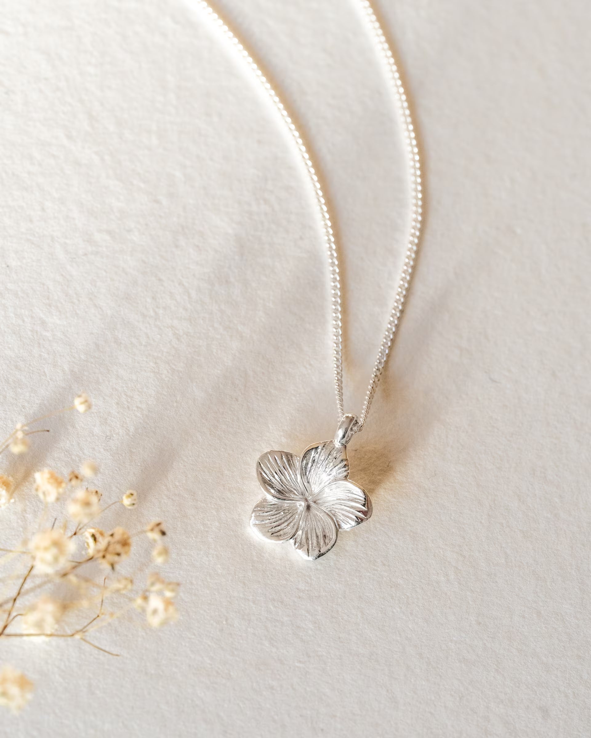 FRANGIPANI FLOWER NECKLACE SILBER, silberne Halskette von ILUH, handgefertigt auf Bali – Elegantes Produktfoto im zarten Boho-Look von einer feinen Halskette aus 925er Silber mit einem detailgetreu strukturierten Frangipani-Blütenanhänger, flach präsentiert auf einem hellen Büttenpapier als Untergrund neben feinen Trockenblumen.
