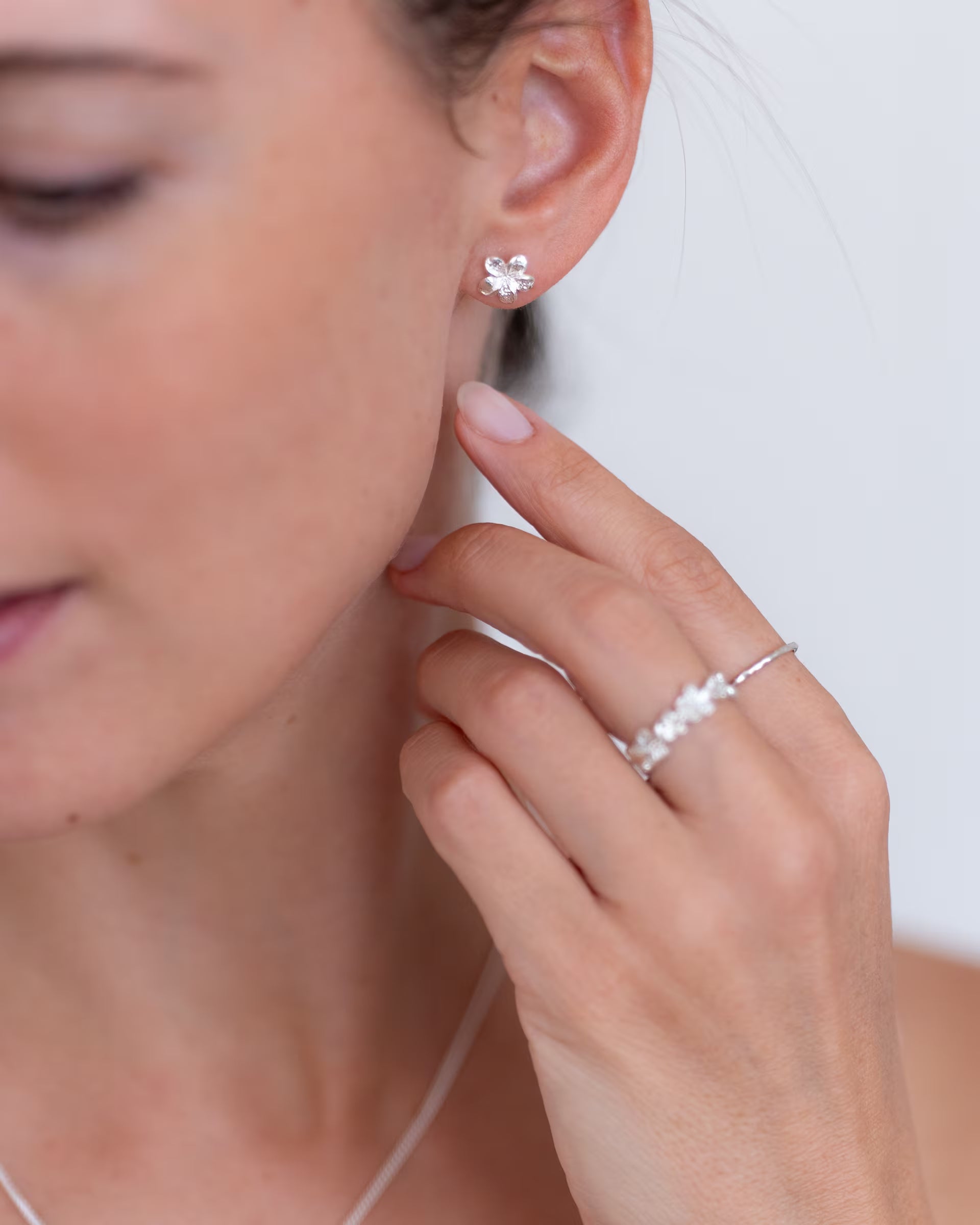 Nahaufnahme im minimalistisch-eleganten Look von einer Frau im Profil, die eine Hand sanft an die Wange legt und eine Kombination aus auf Bali handgefertigtem silbernem Schmuck – bestehend aus den zarten FRANGIPANI OHRSTECKERN, dem detailreichen FRANGIPANI FLOWER RING aus aneinandergereihten Blüten sowie dem feinen, gehämmerten SARI STACKING RING – aus 925er Silber von der Schmuckmarke ILUH trägt.