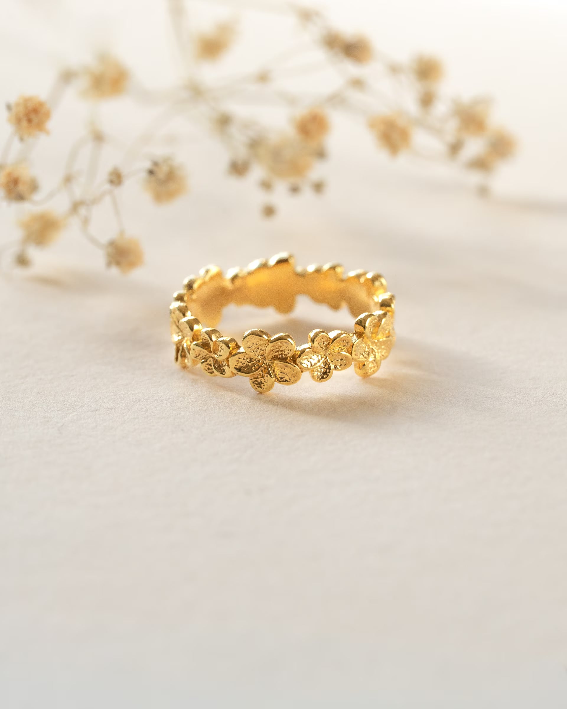 FRANGIPANI FLOWER RING GOLD, goldener Ring von ILUH, handgefertigt auf Bali – Elegantes Produktfoto im zarten Boho-Look von einem filigranen Ring aus 18k vergoldetem 925er Silber, der aus einer Reihe rundherum aneinandergereihter, detailgetreu strukturierter Frangipani-Blüten besteht, präsentiert auf einem hellen Untergrund vor dezenten Trockenblumen.