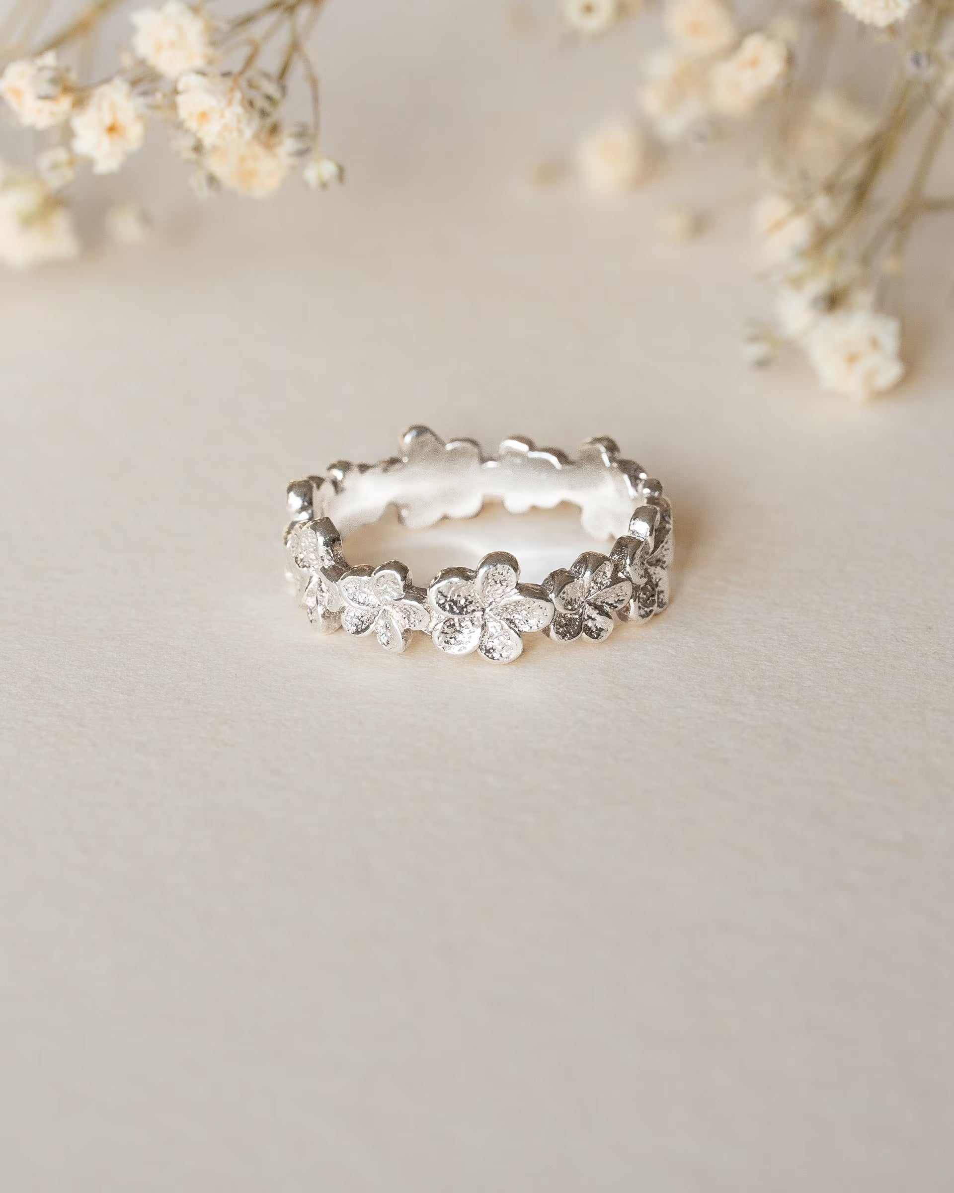 FRANGIPANI FLOWER RING SILBER, silberner Ring von ILUH, handgefertigt auf Bali – Elegantes Produktfoto im zarten Boho-Look von einem filigranen, floralen Ring aus 925er Silber, der aus einer Reihe rundherum aneinandergereihter, detailgetreu strukturierter Frangipani-Blüten besteht, präsentiert auf einem hellen Untergrund vor dezenten Trockenblumen.