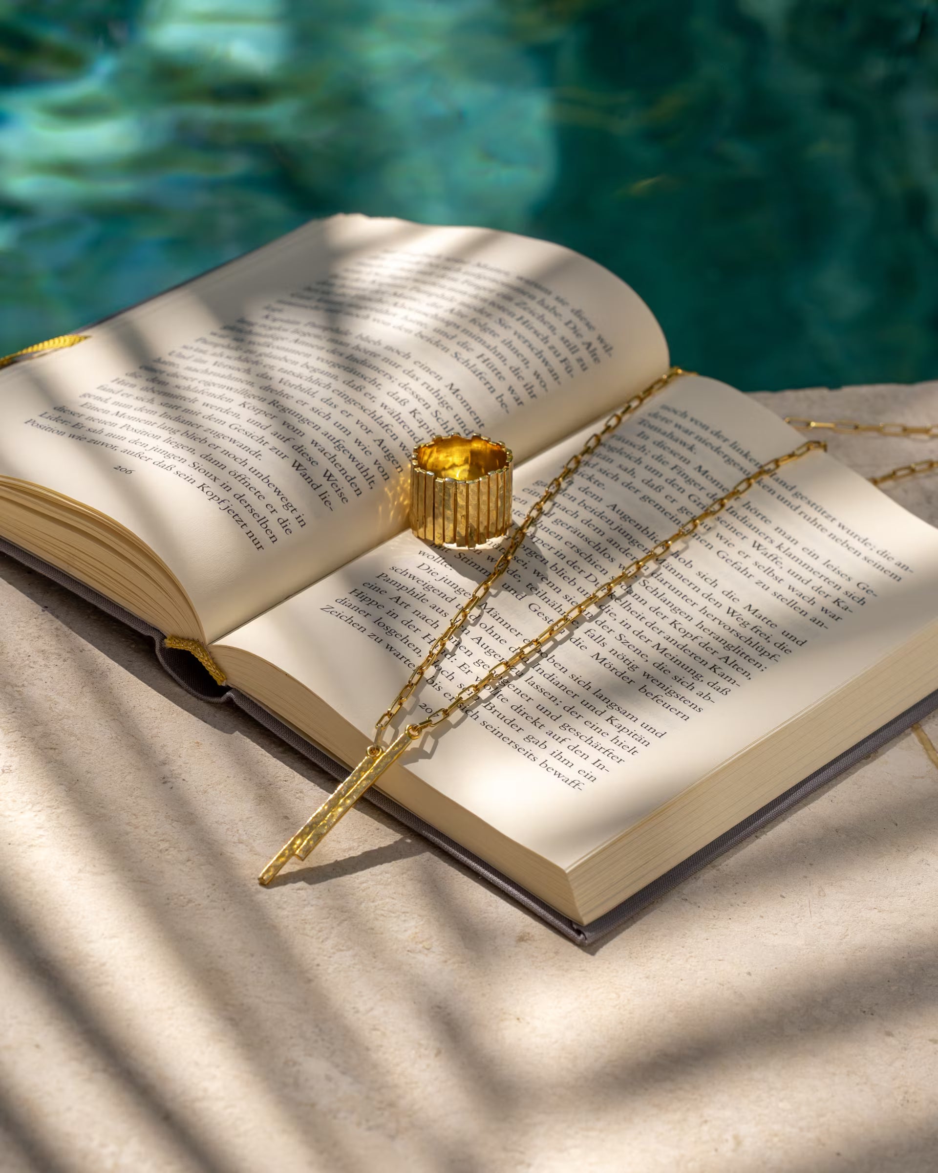 Produktfoto im sommerlichen Stil von der auf Bali handgefertigten goldenen FUSE Paperchain-Kette mit gehämmertem Stab-Anhänger und einem breiten, strukturierten FUSE Statement-Ring aus 18k vergoldetem 925er Silber von der Schmuckmarke ILUH, kunstvoll platziert auf einem geöffneten Buch am Poolrand mit Schatten von Palmenblättern.