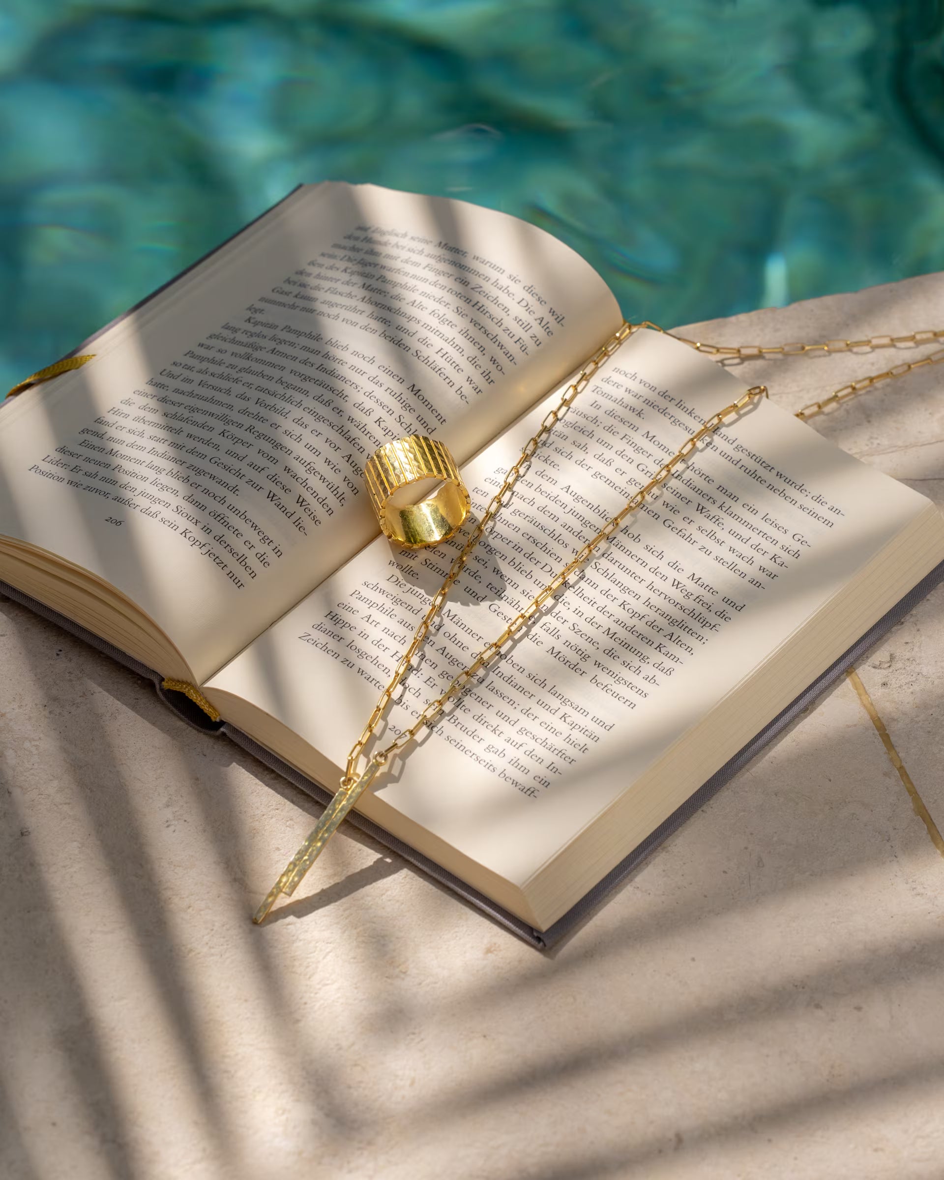 Produktfoto im sommerlichen Stil von der auf Bali handgefertigten goldenen FUSE Paperchain Kette mit strukturiertem Stab-Anhänger und dem breiten FUSE Statement-Ring aus 18k vergoldetem 925er Silber von der Schmuckmarke ILUH, dekorativ platziert auf den Seiten eines geöffneten Buches am Poolrand mit Palmenschatten.
