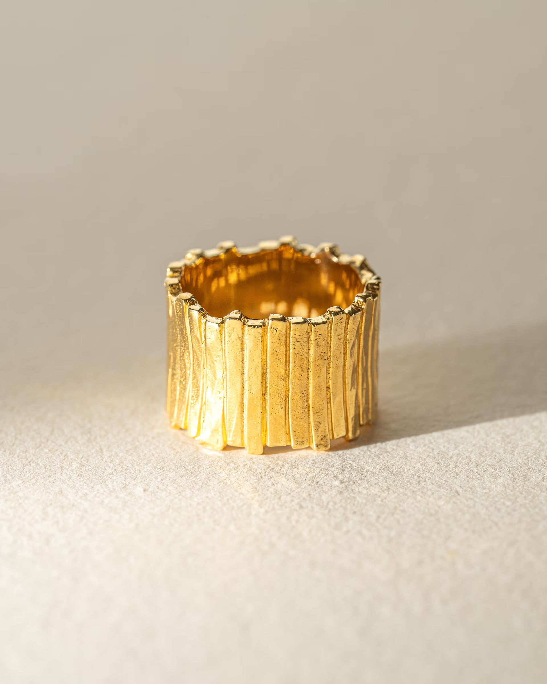 FUSE STATEMENT RING GOLD, breiter goldener Ring von ILUH, handgefertigt auf Bali – Elegantes Produktfoto im minimalistischen Boho-Look von einem sehr breiten, markanten Statement-Ring aus 18k vergoldetem 925er Silber mit einer stark strukturierten, gehämmerten Oberfläche im organischen Stab-Design, effektvoll präsentiert im hellen Streiflicht auf neutralem, beigem Untergrund.