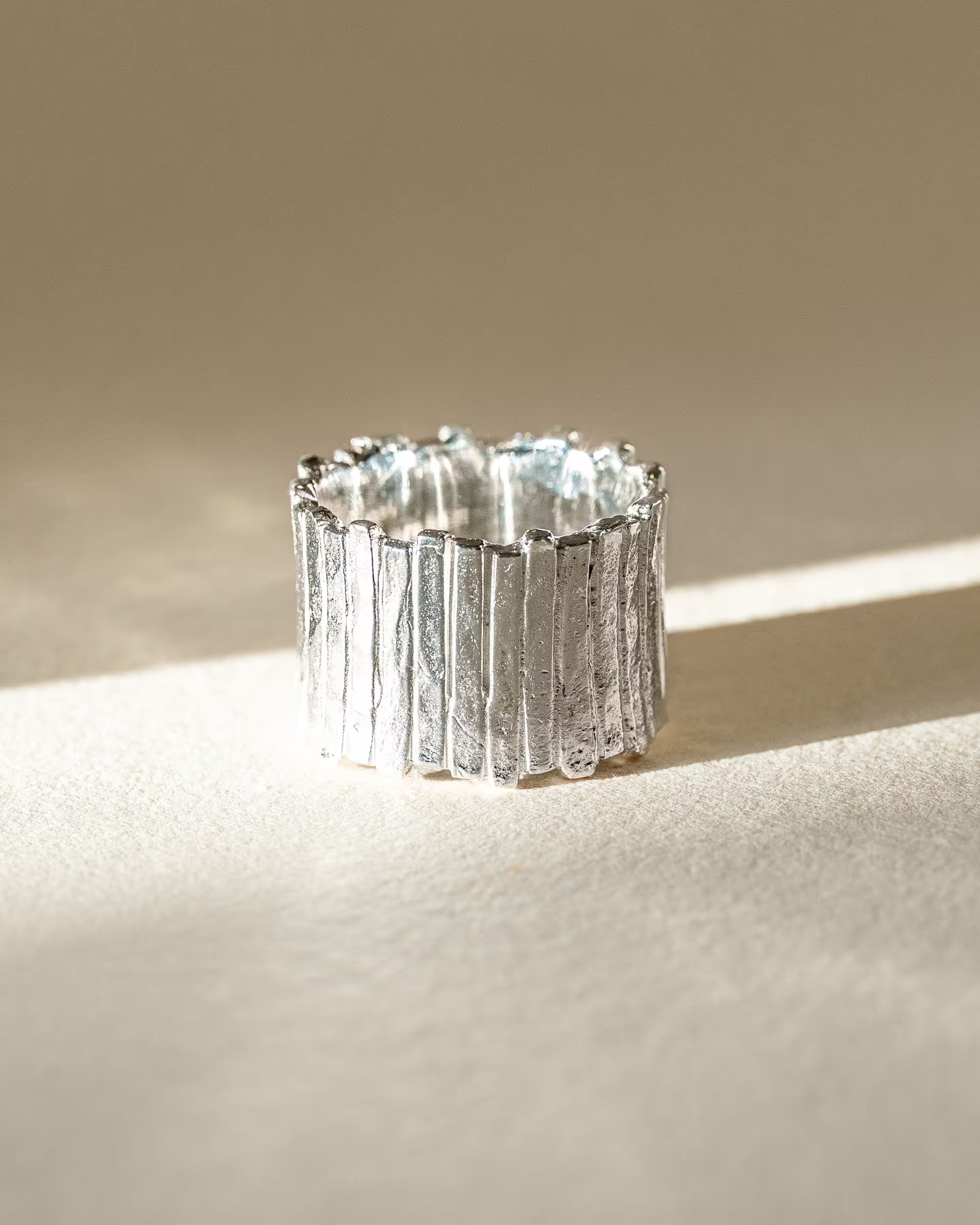 FUSE STATEMENT RING SILBER, breiter silberner Ring von ILUH, handgefertigt auf Bali – Elegantes Produktfoto im minimalistischen Boho-Look von einem sehr breiten,  markanten Statement-Ring aus 925er Silber mit einer stark strukturierten, gehämmerten Oberfläche im organischen Stab-Design, effektvoll präsentiert im hellen Streiflicht auf neutralem, beigem Untergrund.
