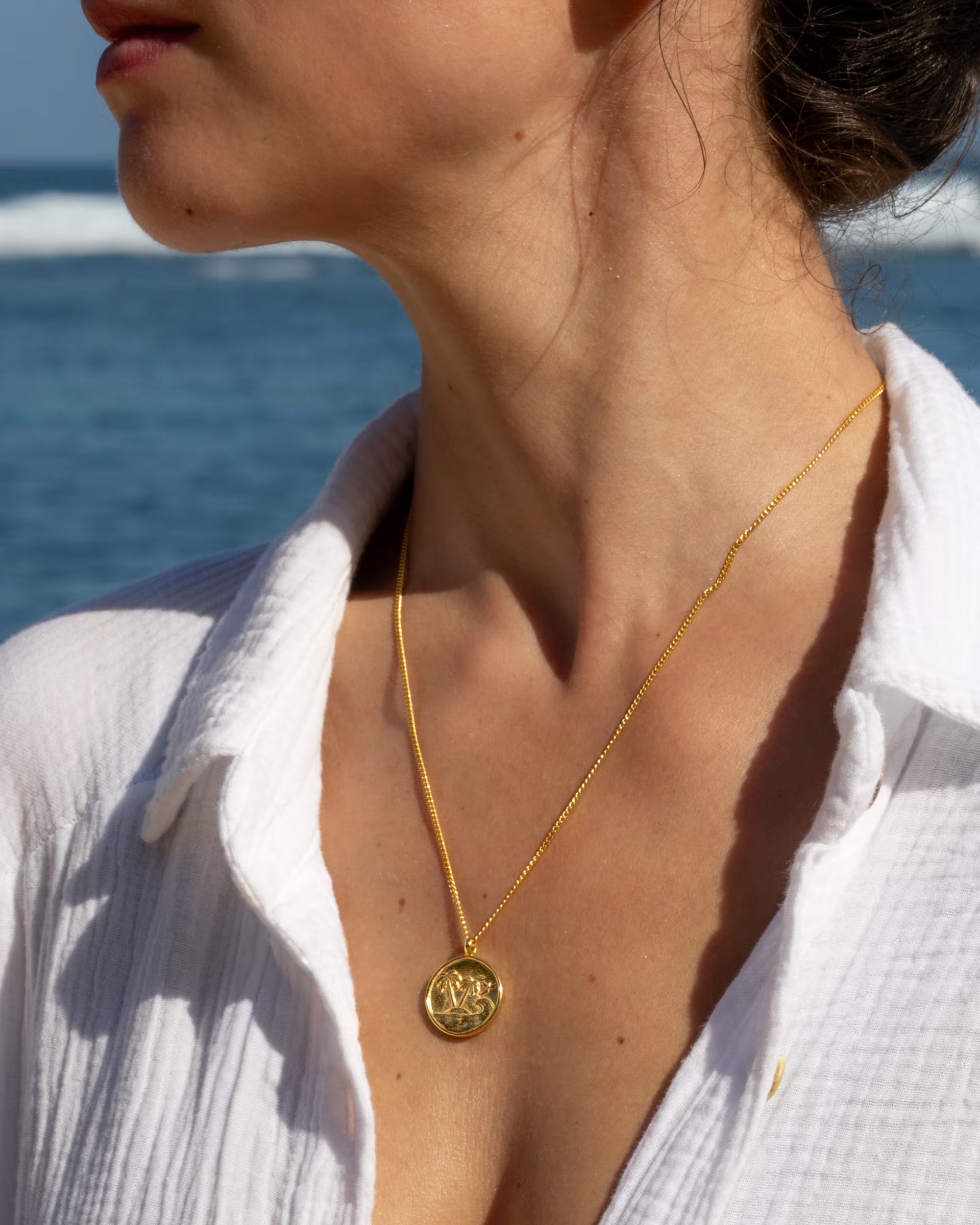 Nahaufnahme im sommerlichen Beach-Look von einer Frau in einer weißen Musselin-Bluse, die die auf Bali handgefertigte goldene GILI PARADISE ISLAND Kette mit einem runden Münz-Anhänger mit Palmen-Motiv aus 18k vergoldetem 925er Silber von der Schmuckmarke ILUH am Meer trägt.