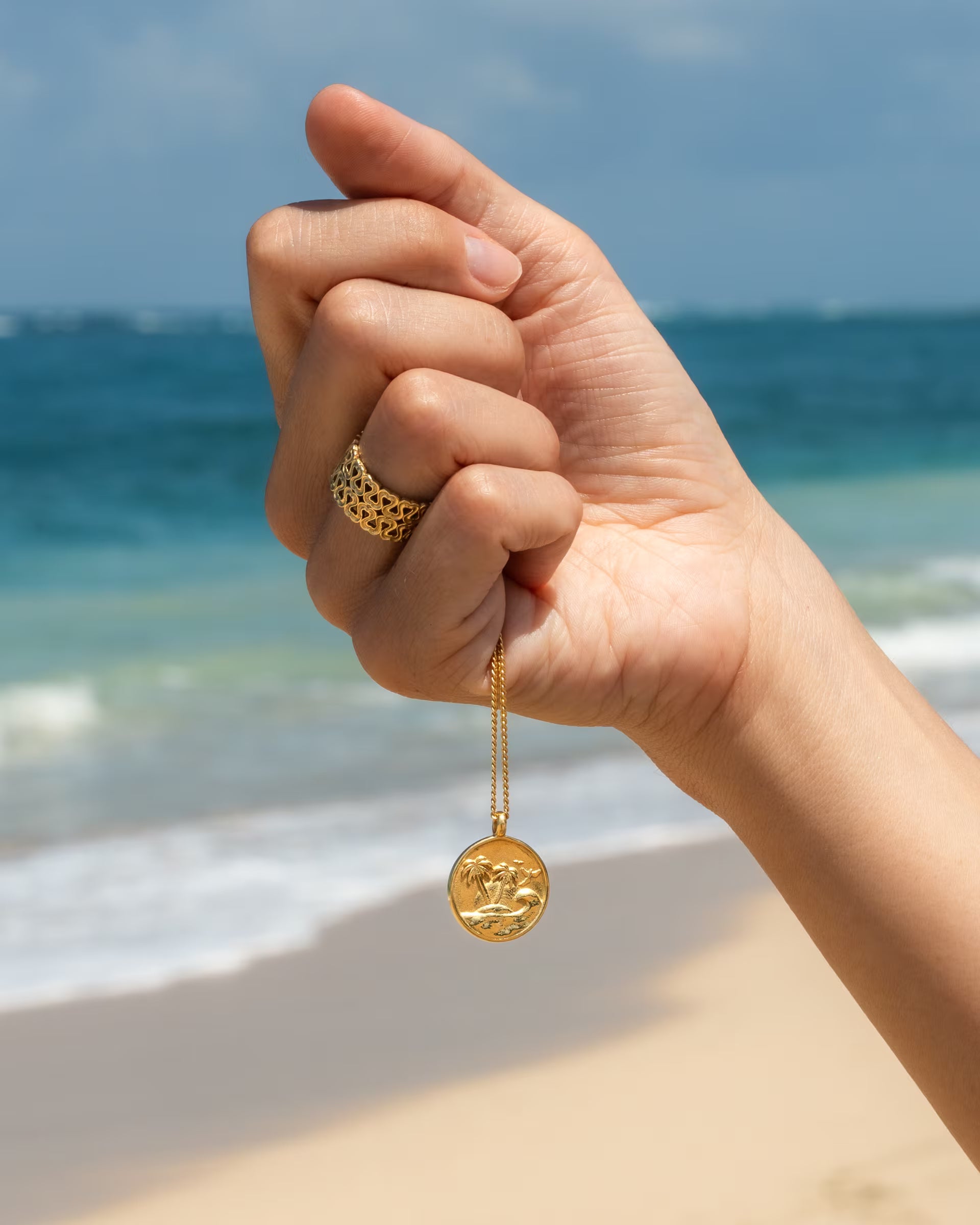 Stimmungsvolle Nahaufnahme im maritimen Beach-Look von einer angehobenen Hand am Sandstrand vor dem blauen Meer, die die auf Bali handgefertigte goldene GILI PARADISE ISLAND KETTE mit einem runden, geprägten Insel- und Palmen-Münzanhänger herabhängen lässt und den breiten, offenen ENDLESS LOVE RING mit Herz-Muster aus 18k vergoldetem 925er Silber von der Schmuckmarke ILUH trägt.