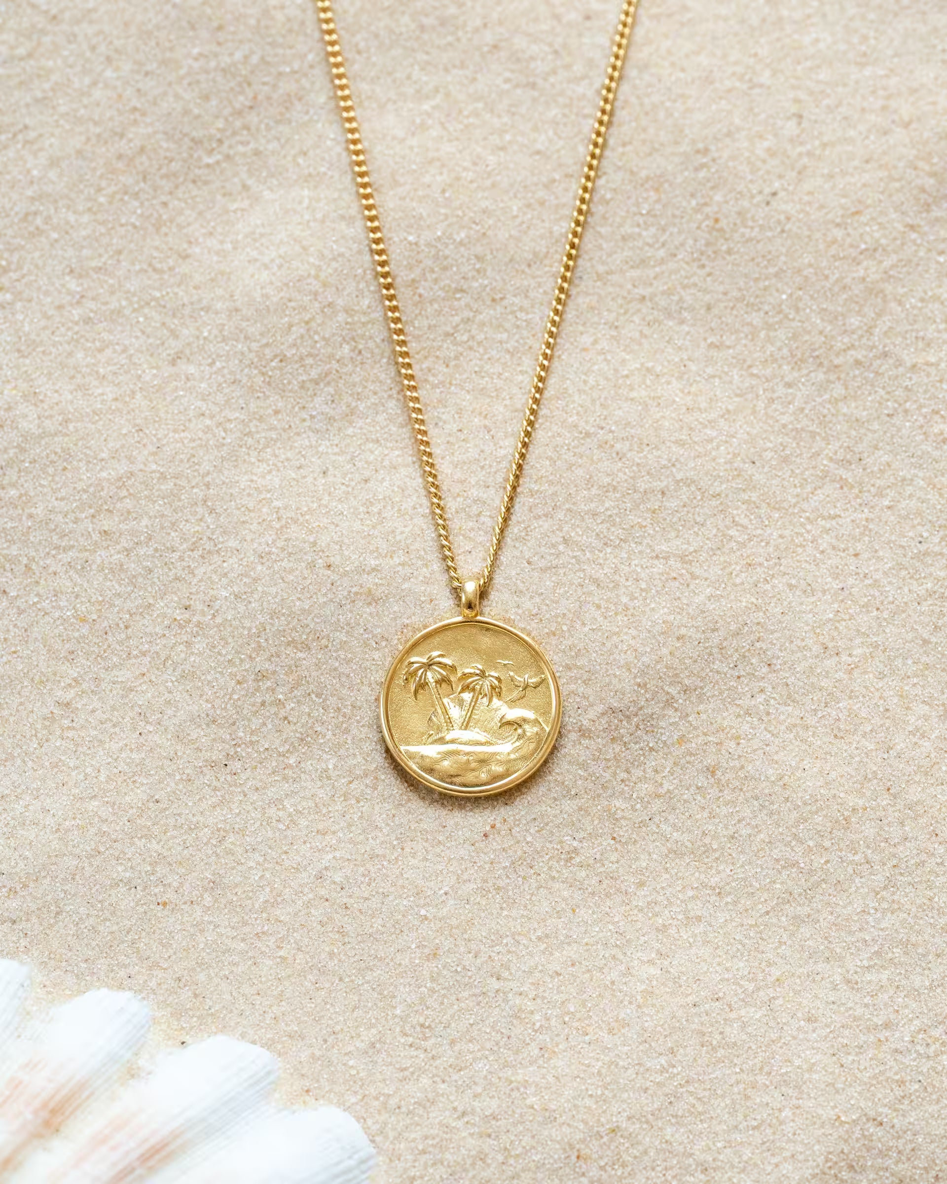 GILI PARADISE ISLAND NECKLACE GOLD, goldene Halskette von ILUH, handgefertigt auf Bali – Elegantes Produktfoto im maritimen Beach-Look von einer feinen Halskette aus 18k vergoldetem 925er Silber mit einem runden Münzanhänger, auf dem ein detailliertes Insel-Motiv mit Palmen und Wellen eingeprägt ist, flach präsentiert im feinen Sandstrand neben dem Ansatz einer hellen Muschel.