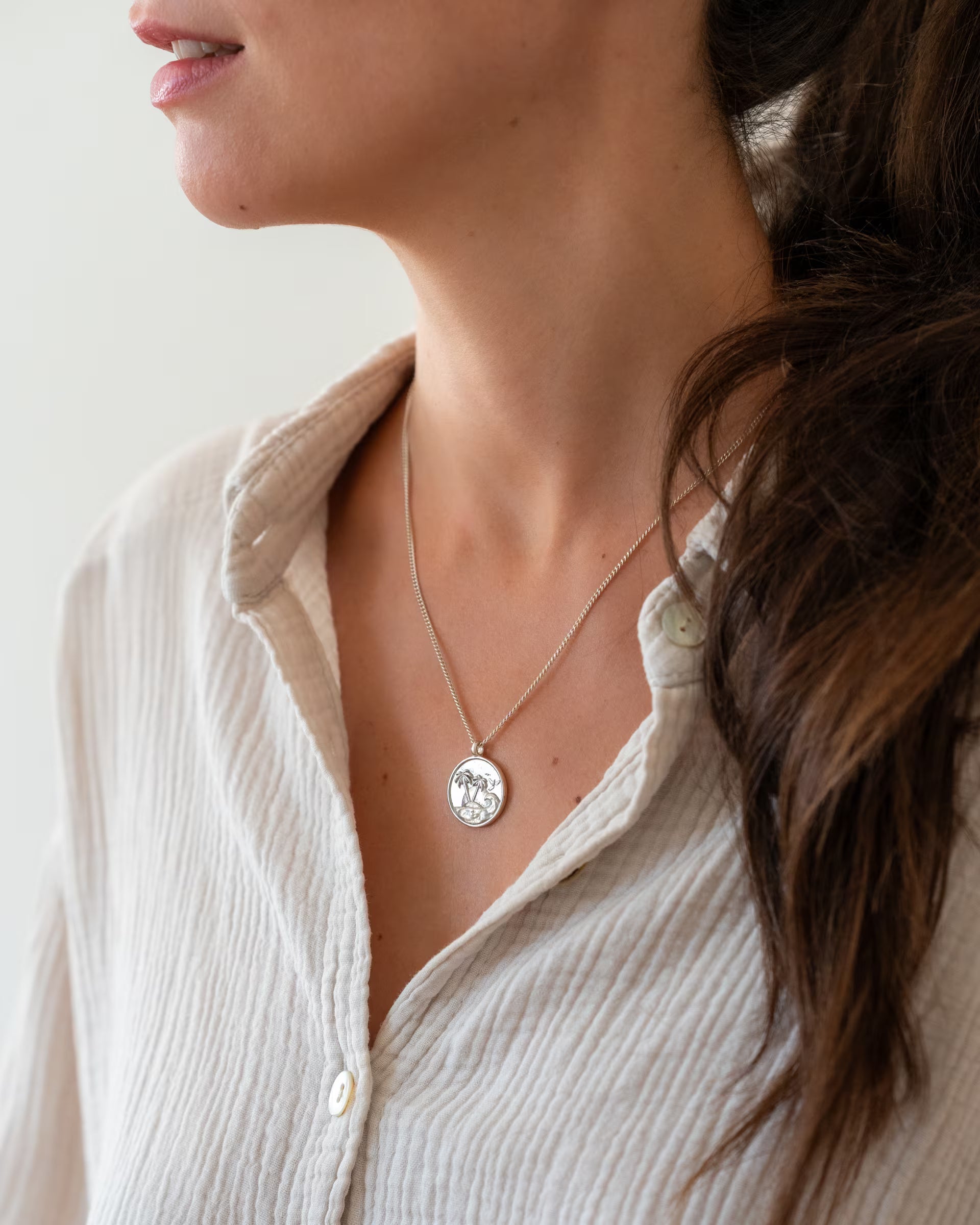 Nahaufnahme im minimalistischen Look von einer Frau in einer weißen Musselin Bluse, die die auf Bali handgefertigte silberne GILI PARADISE ISLAND Kette mit einem runden Münz-Anhänger mit Palmen-Motiv aus 925er Silber von der Schmuckmarke ILUH trägt.