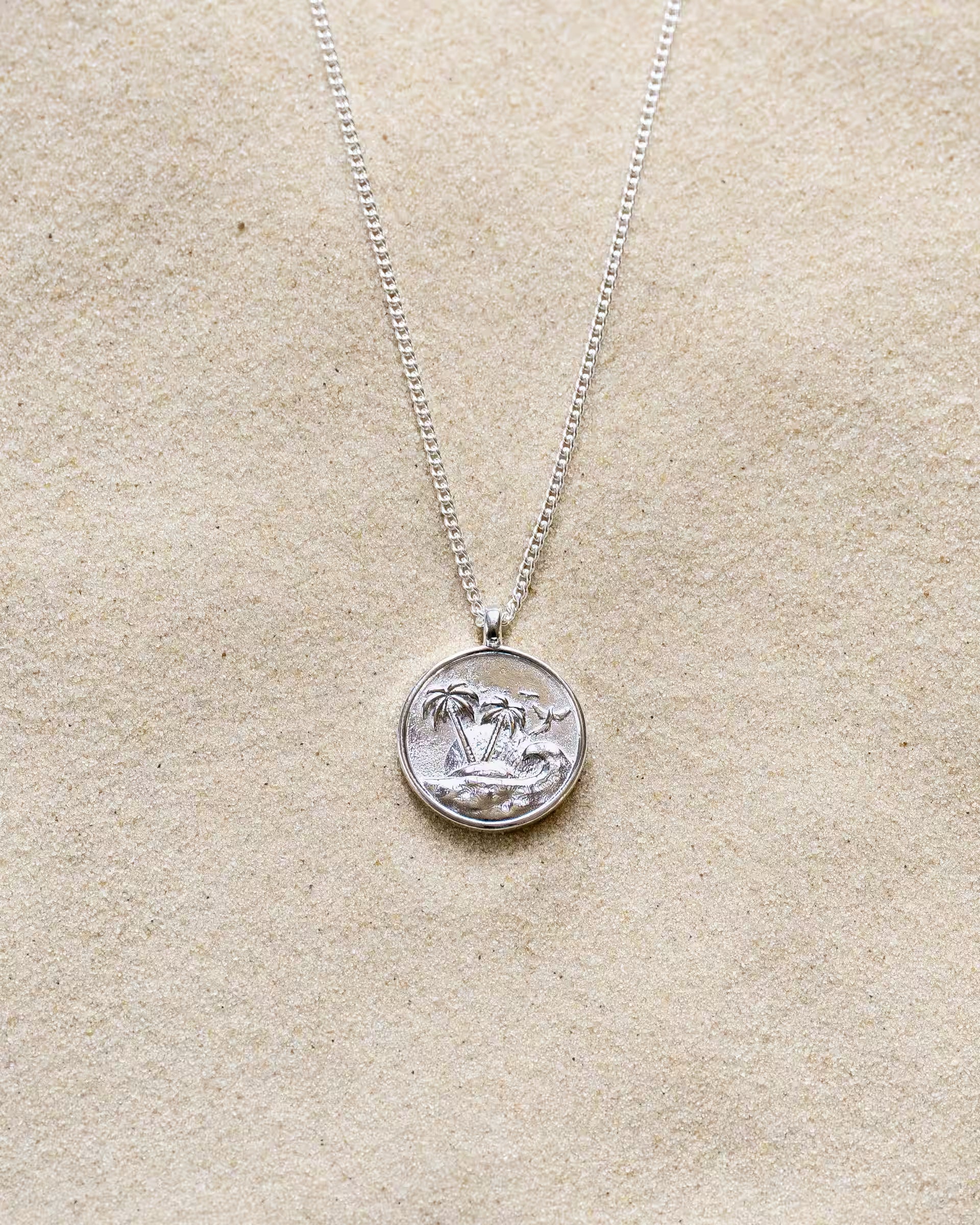 GILI PARADISE ISLAND NECKLACE SILBER, silberne Halskette von ILUH, handgefertigt auf Bali – Elegantes Produktfoto im maritimen Beach-Look von einer feinen Halskette aus 925er Silber mit einem runden Münzanhänger, auf dem ein detailliertes Insel-Motiv mit Palmen und Wellen eingeprägt ist, flach präsentiert im feinen Sandstrand.