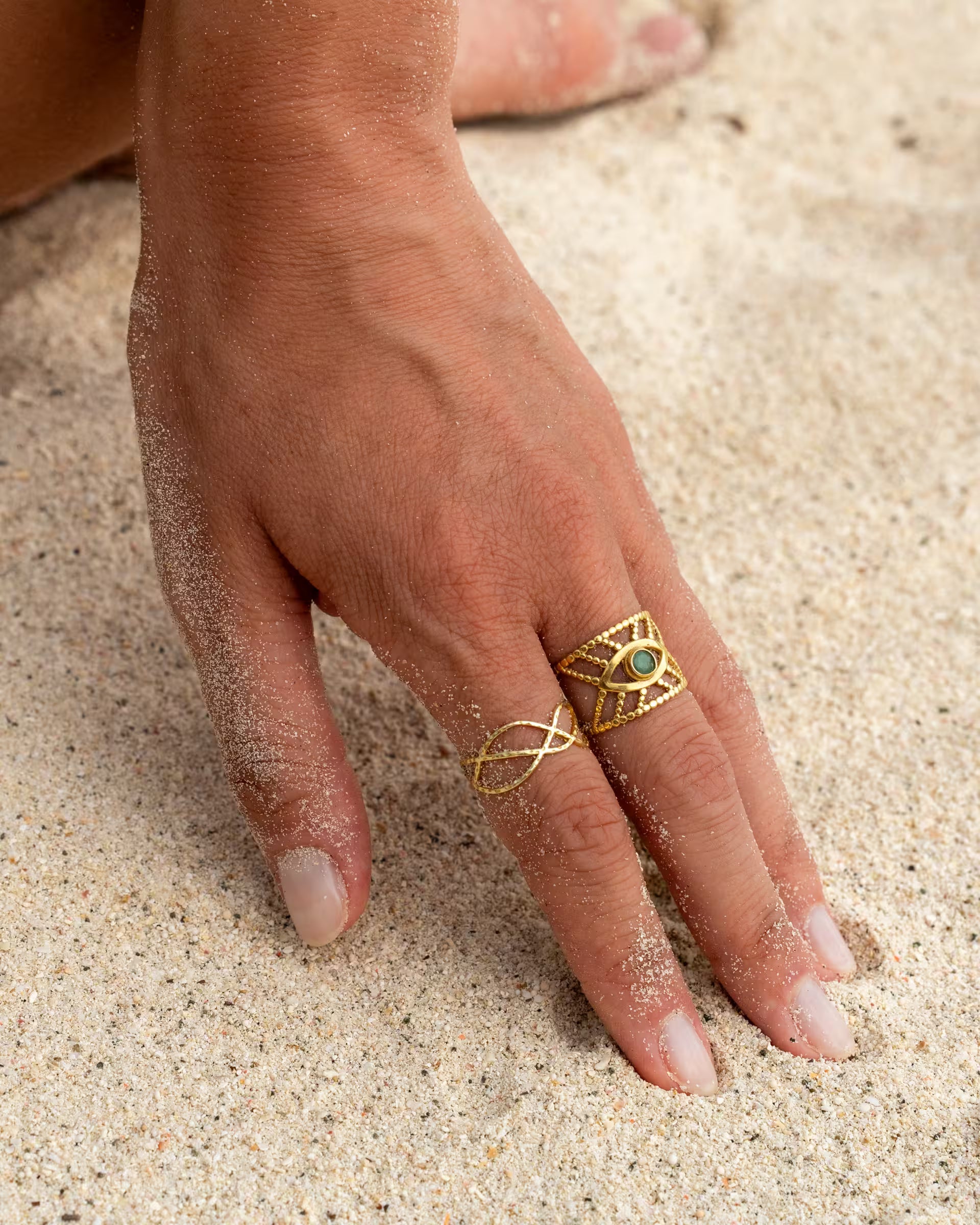 Nahaufnahme von dem handgefertigten INTUITION Ring mit grünem Smaragd-Edelstein und LOTUS Ring aus 18k vergoldetem 925er Silber von der Schmuckmarke ILUH, getragen von einer jungen Frau im Stacking-Look an einer Hand im hellen Sand am Strand.