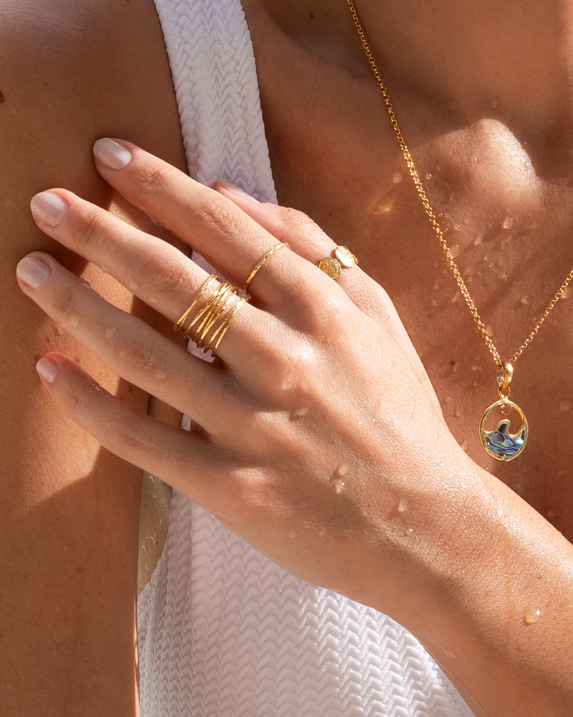 Nahaufnahme im sommerlichen Beach-Look von einer Hand mit Wassertropfen auf der Haut, die eine Kombination aus auf Bali handgefertigten goldenen Ringen – darunter den AURA Ring im Stacking-Look, den feinen SARI Ring und den CHANDRA MOONPHASES Ring – sowie die passende LAJU Wave Abalone Kette aus 18k vergoldetem 925er Silber von der Schmuckmarke ILUH trägt.