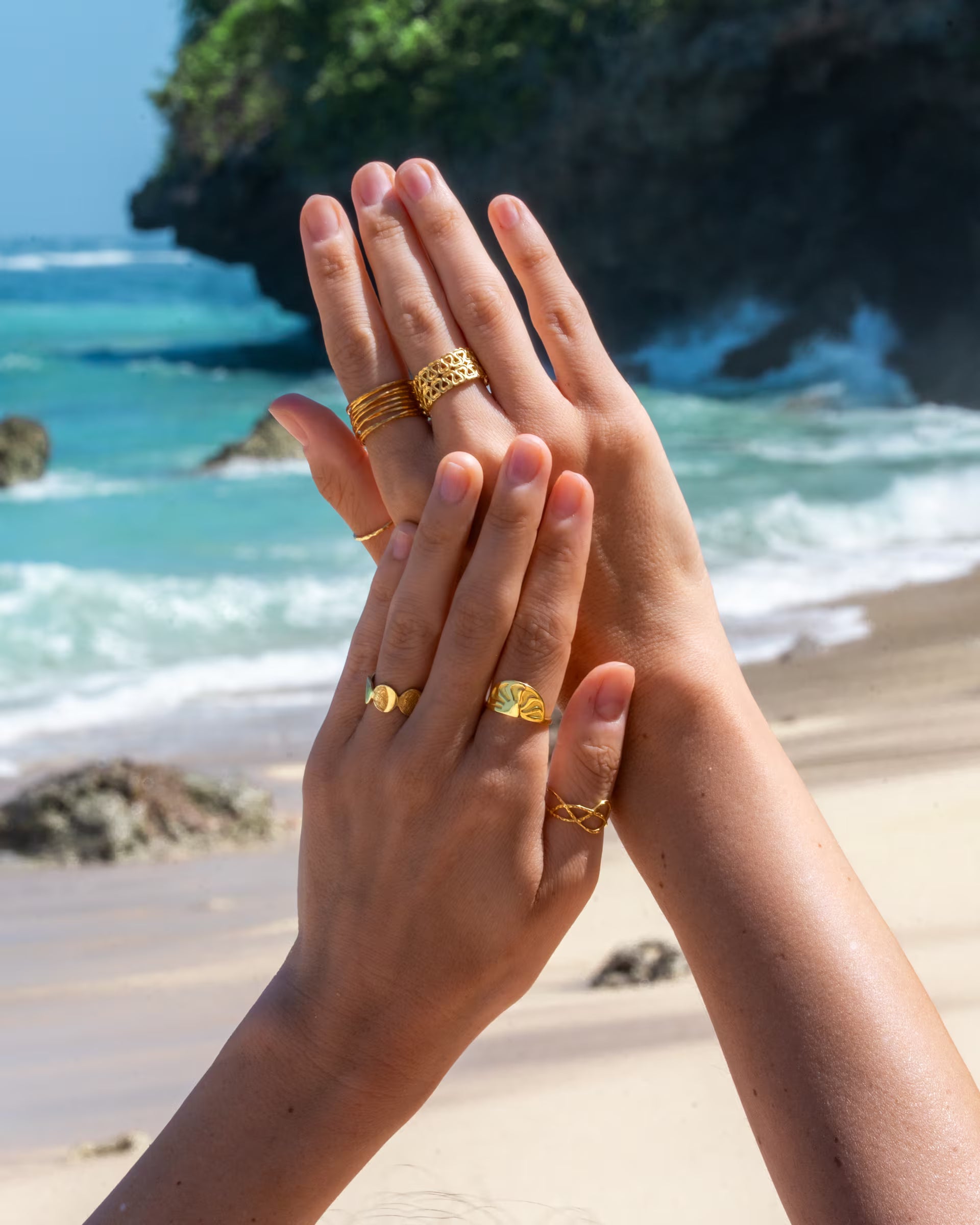 Stimmungsvolle Nahaufnahme im sommerlichen Beach-Look von zwei elegant emporgehobenen Händen an einem tropischen Strand mit türkisblauem Meer und Felsen im Hintergrund, die eine Kombination aus auf Bali handgefertigtem goldenem Schmuck – darunter den ARUNA SUNRISE Ring, den AURA RING im Stacking-Look, den offenen ENDLESS LOVE RING mit Herz-Muster sowie den geschwungenen LOTUS RING – aus 18k vergoldetem 925er Silber von der Schmuckmarke ILUH tragen.