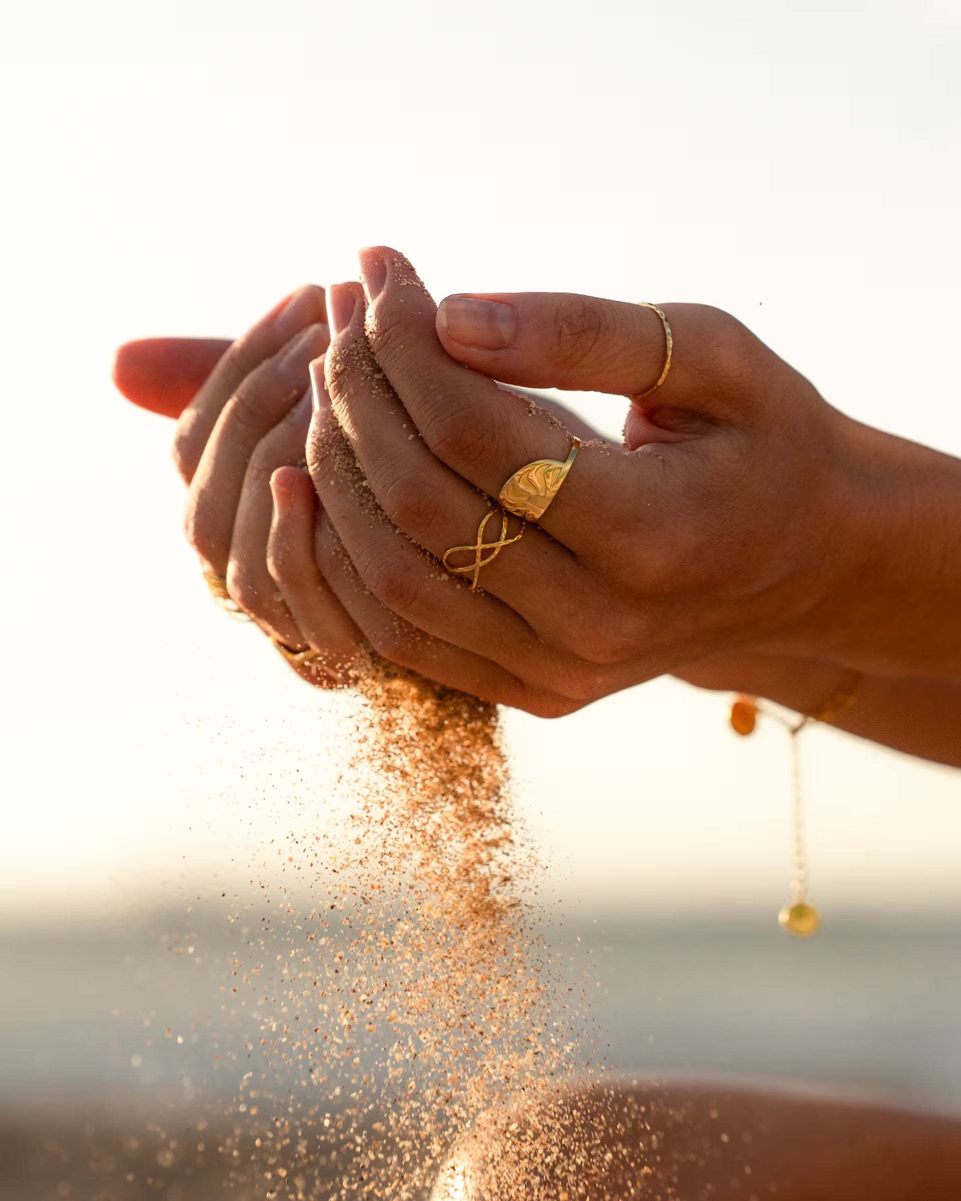 Stimmungsvolle Nahaufnahme im sommerlichen Beach-Look von zwei Händen im Gegenlicht des Sonnenuntergangs, durch die feiner Strandsand rinnt, während sie eine Kombination aus auf Bali handgefertigtem goldenem Schmuck – darunter den ARUNA SUNRISE Ring mit Sonnen-Motiv, den geschwungenen LOTUS RING, den feinen, gehämmerten SARI STACKING RING sowie das zarte CHAKRA ARMBAND – aus 18k vergoldetem 925er Silber von der Schmuckmarke ILUH tragen.