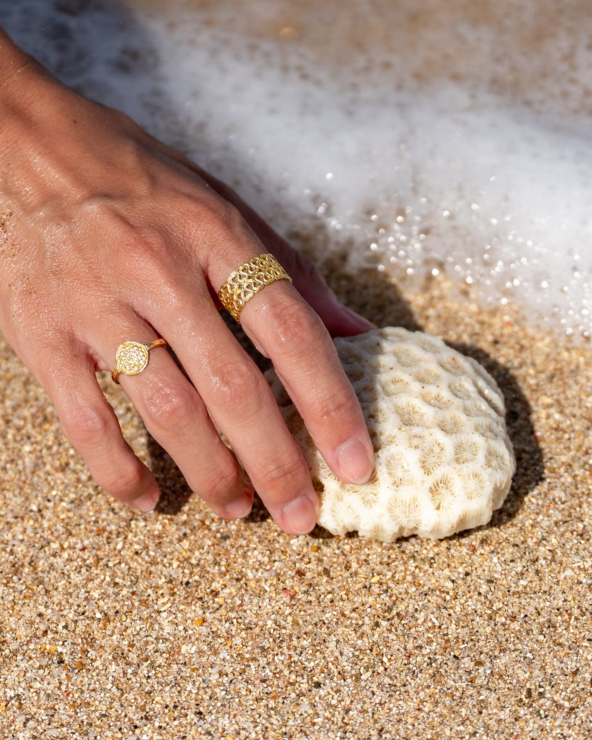 Handgefertigter goldener ENDLESS LOVE Ring und Chakra-Motivring aus 18k vergoldetem 925er Silber von der Schmuckmarke ILUH, getragen an der Hand einer jungen Frau im Sand, die eine weiße Koralle am Strand berührt, während eine Welle mit weißer Gischt heranspült.