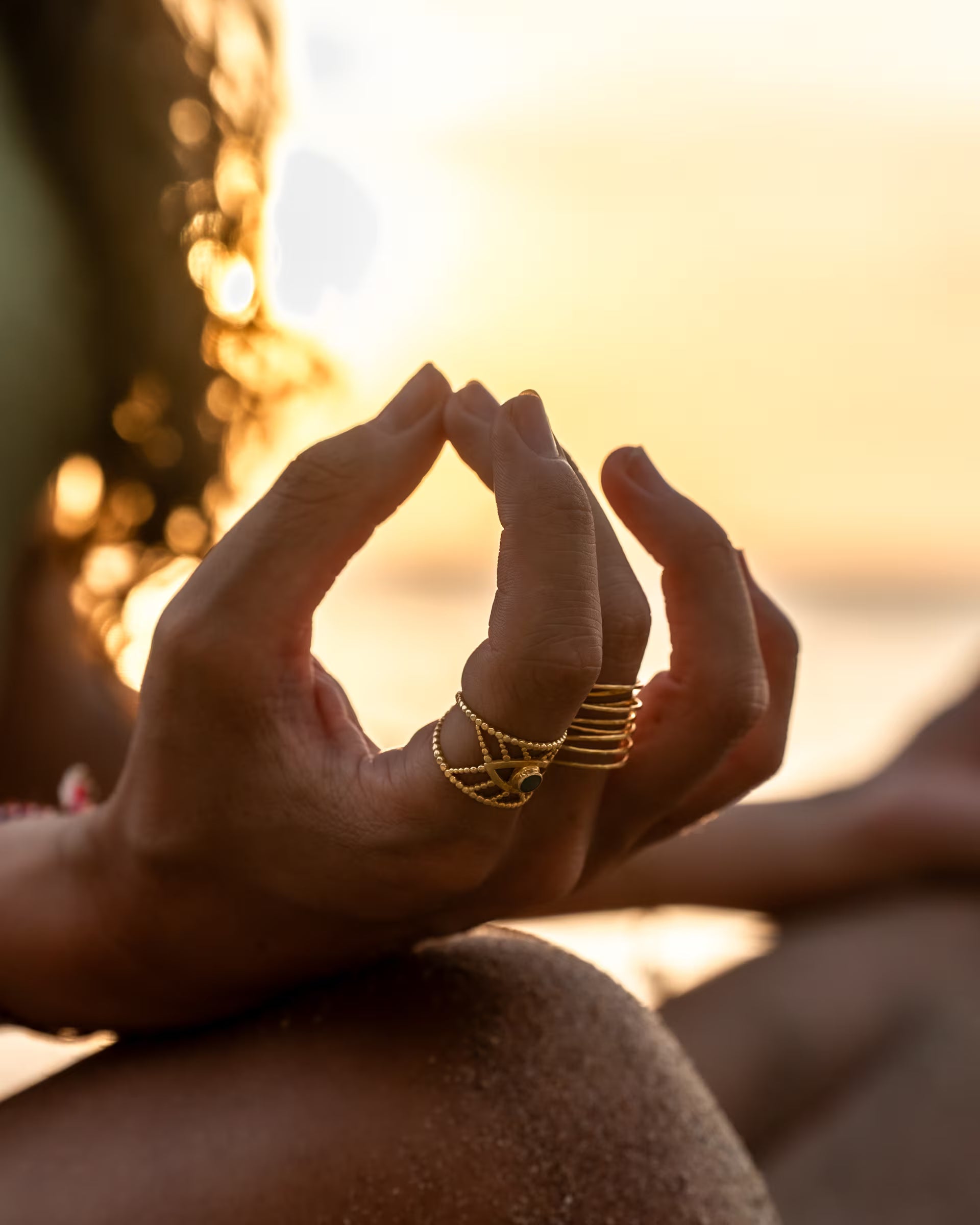 Stimmungsvolle Nahaufnahme im spirituellen Yoga-Stil von einer Hand in einer Meditations-Geste (Mudra) am Strand, die eine Kombination aus auf Bali handgefertigtem goldenem Schmuck – darunter den breiten, offenen INTUITION RING mit grünem Edelstein sowie den gehämmerten AURA RING im Stacking-Look – aus 18k vergoldetem 925er Silber von der Schmuckmarke ILUH im warmen, goldenen Licht des Sonnenuntergangs trägt.