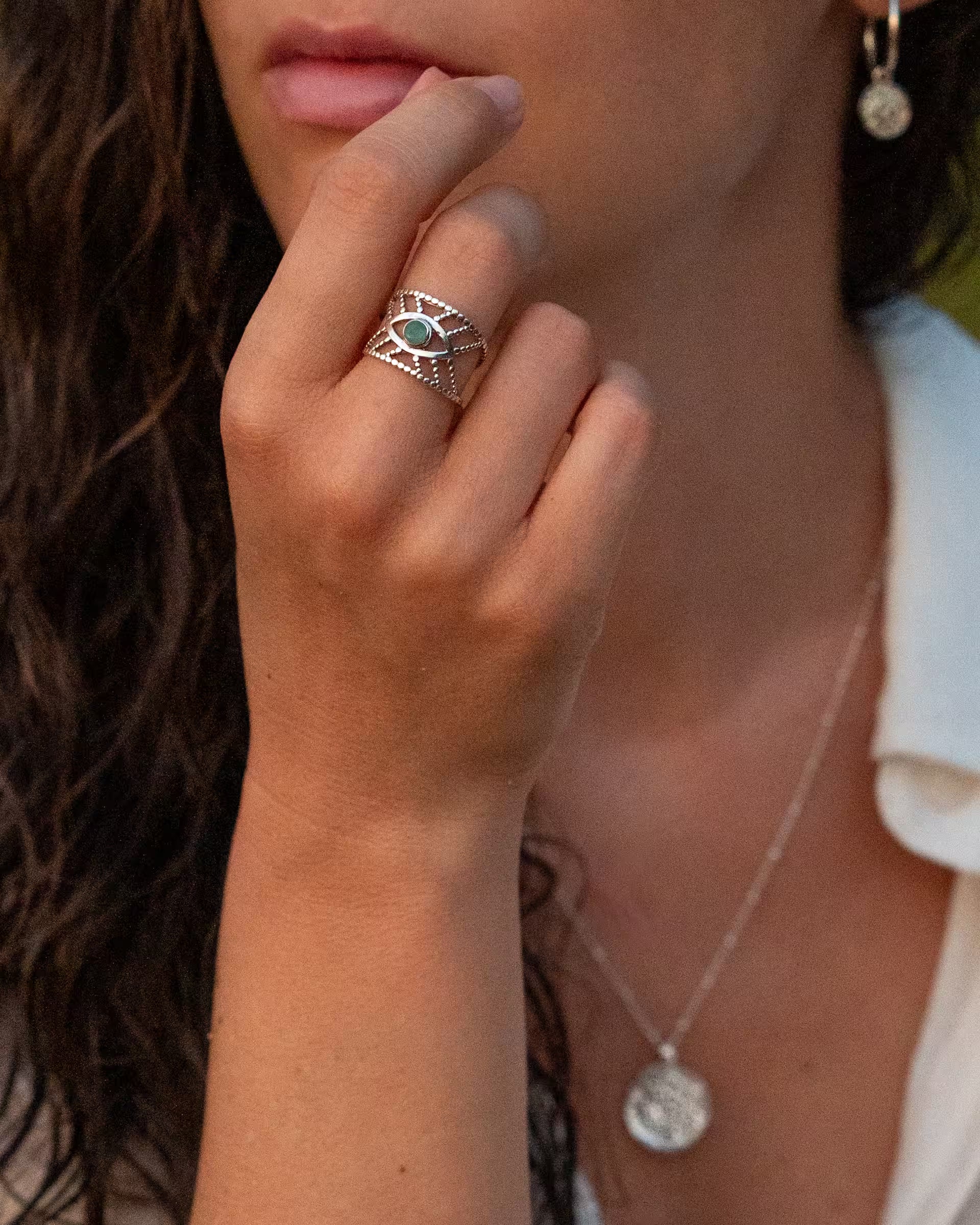 Handgefertigter silberner Intuition Ring mit grünem Smaragd-Edelstein aus 925er Silber von der Schmuckmarke ILUH, getragen von einer jungen Frau zum weißen Hemd im Beach-Look auf Bali, kombiniert mit einer Chakra-Kette und Chakra Creolen-Ohrringen, während ihre Hand sanft ihre Lippen berührt.
