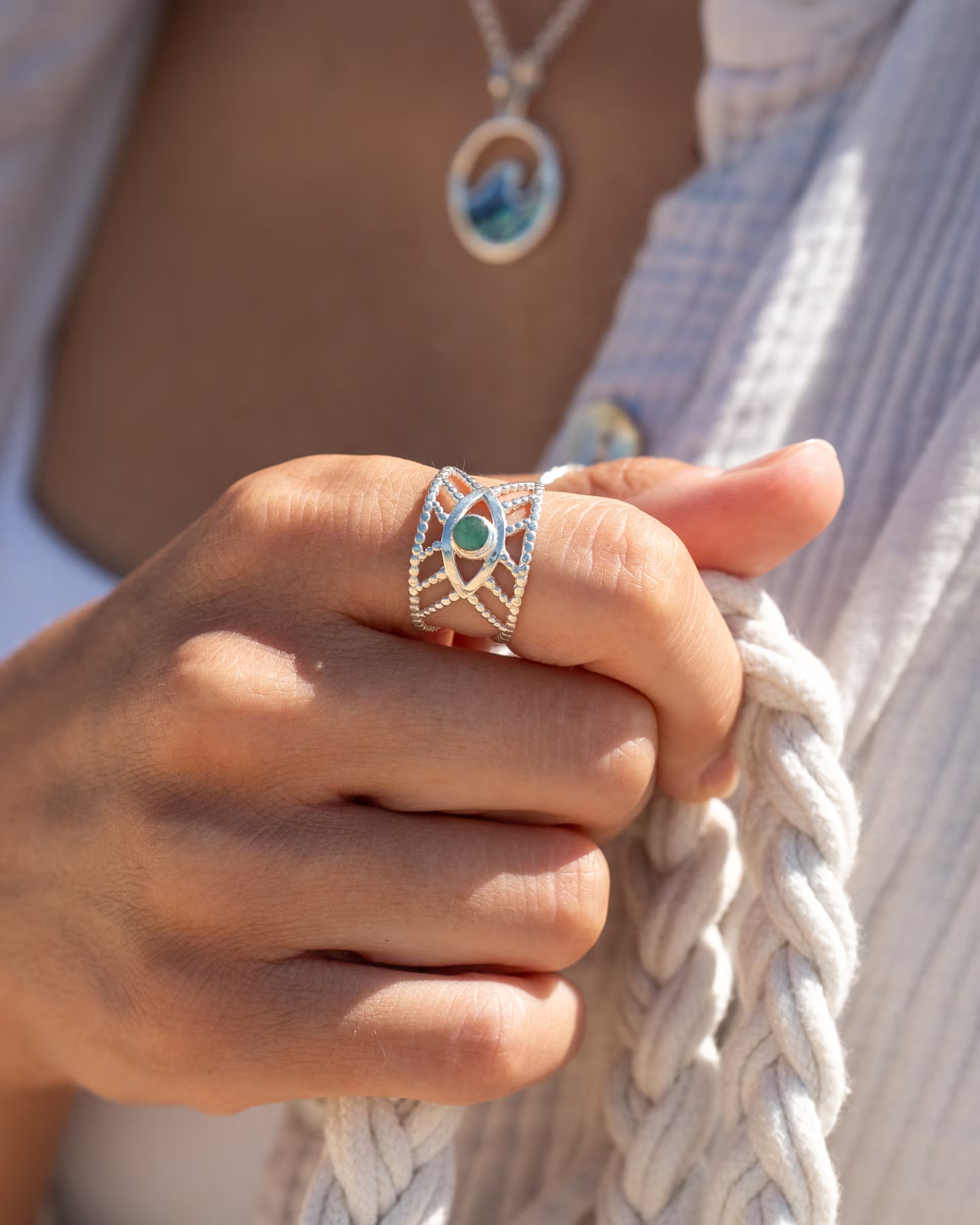 Nahaufnahme im sommerlichen Boho-Look von einer Frau in einer hellen Musselin-Bluse, die die geflochtenen Henkel einer Tasche hält und eine Kombination aus auf Bali handgefertigtem silbernem Schmuck – darunter den offenen INTUITION RING mit grünem Smaragd Edelstein und die LAJU WAVE ABALONE KETTE mit rundem Wellen-Anhänger – aus 925er Silber von der Schmuckmarke ILUH trägt.