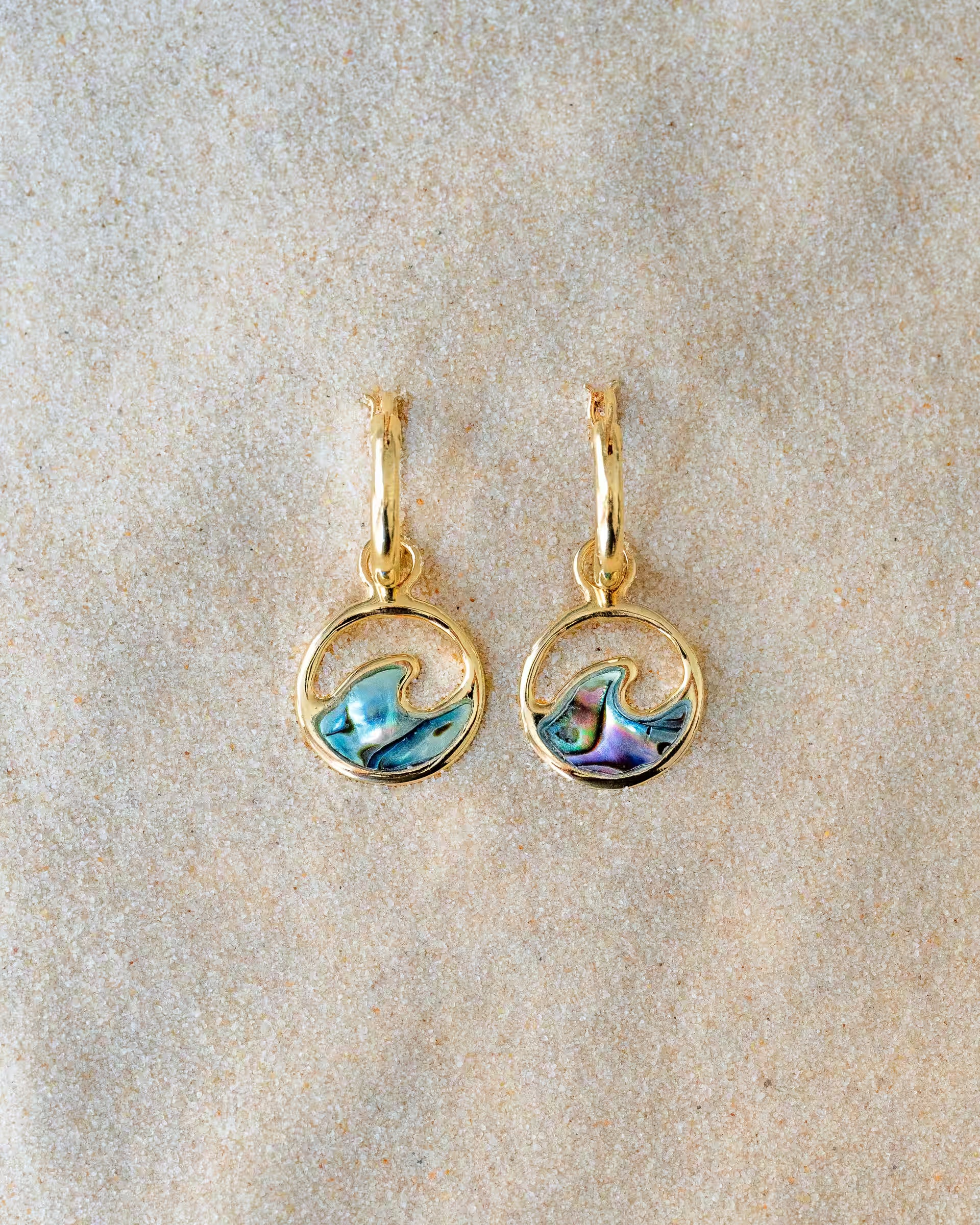 LAJU ABALONE OHRRINGE GOLD, kleine goldene Creolen von ILUH, handgefertigt auf Bali – Elegantes Produktfoto im maritimen Beach-Look von einem Paar kleiner Creolen aus 18k vergoldetem 925er Silber mit runden, offenen Wellen-Anhängern und einer bunt schimmernden Einlage aus echter Abalone-Muschel, flach präsentiert im feinen Sandstrand.