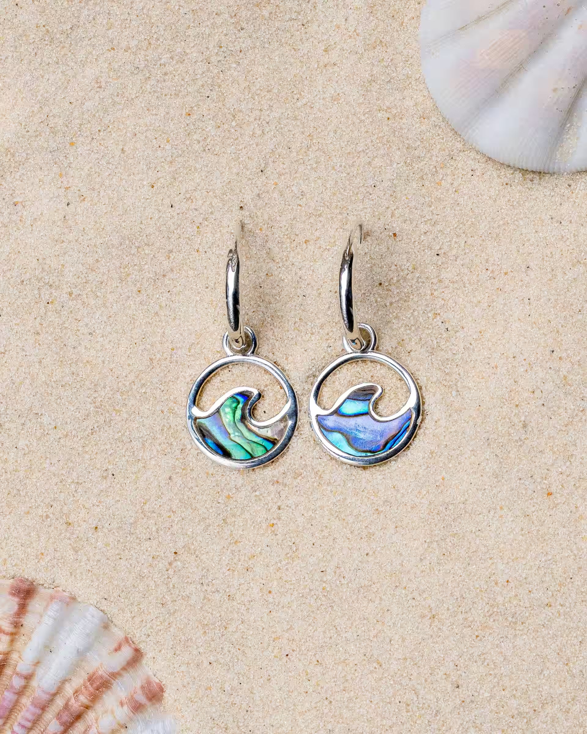 LAJU ABALONE OHRRINGE SILBER, kleine silberne Creolen von ILUH, handgefertigt auf Bali – Elegantes Produktfoto im maritimen Beach-Look von einem Paar kleiner Creolen aus 925er Silber mit runden, offenen Wellen-Anhängern und einer intensiv bunt schimmernden Einlage aus echter Abalone-Muschel, flach präsentiert im feinen Sandstrand zwischen den Ansätzen von zwei hellen Muscheln.