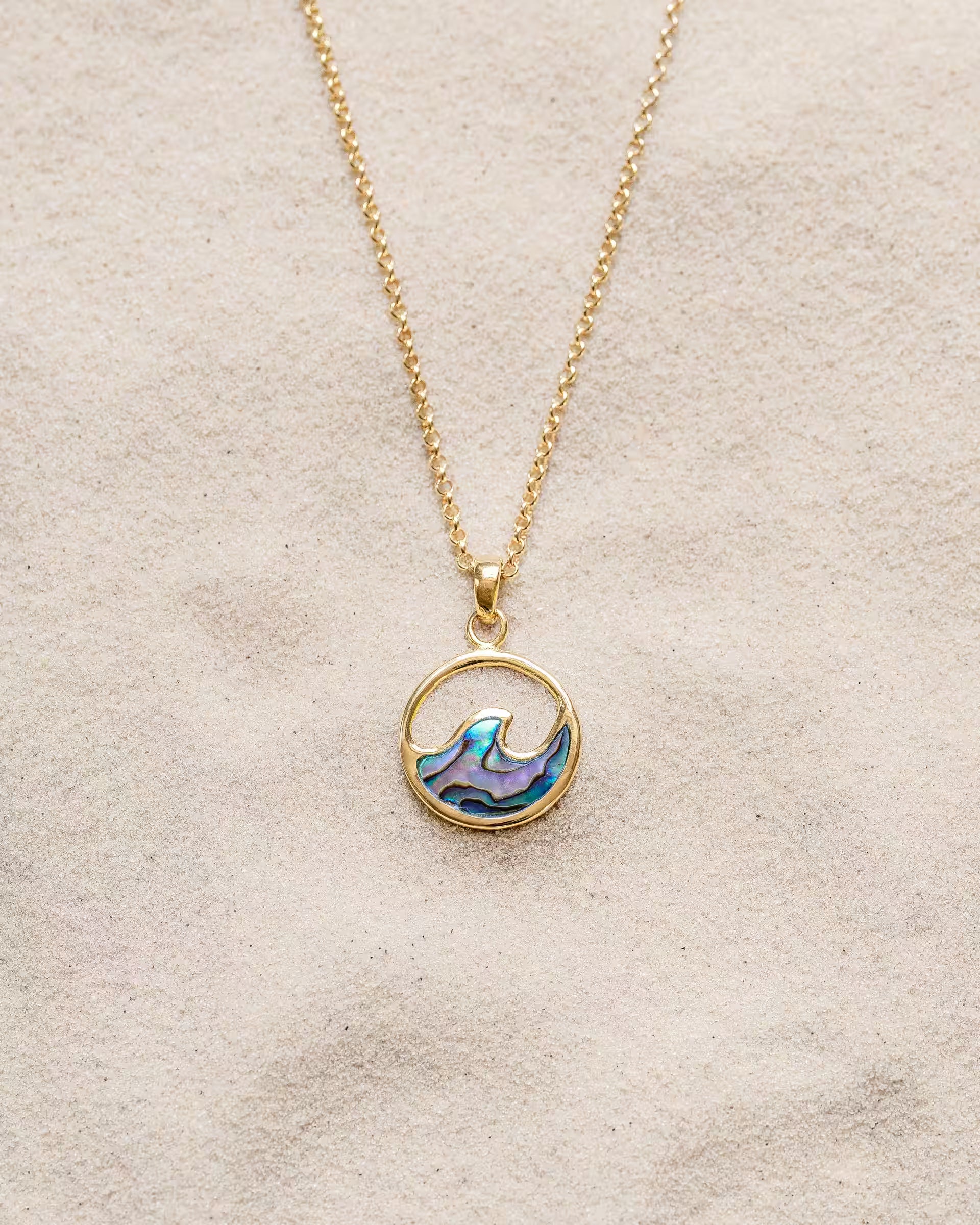 LAJU ABALONE NECKLACE GOLD, goldene Halskette von ILUH, handgefertigt auf Bali – Elegantes Produktfoto im maritimen Beach-Look von einer feinen Halskette aus 18k vergoldetem 925er Silber mit einem runden, offenen Anhänger, der eine geschwungene Meereswelle und eine intensiv blau-violett schimmernde Einlage aus echter Abalone-Muschel zeigt, flach präsentiert im feinen Sandstrand.