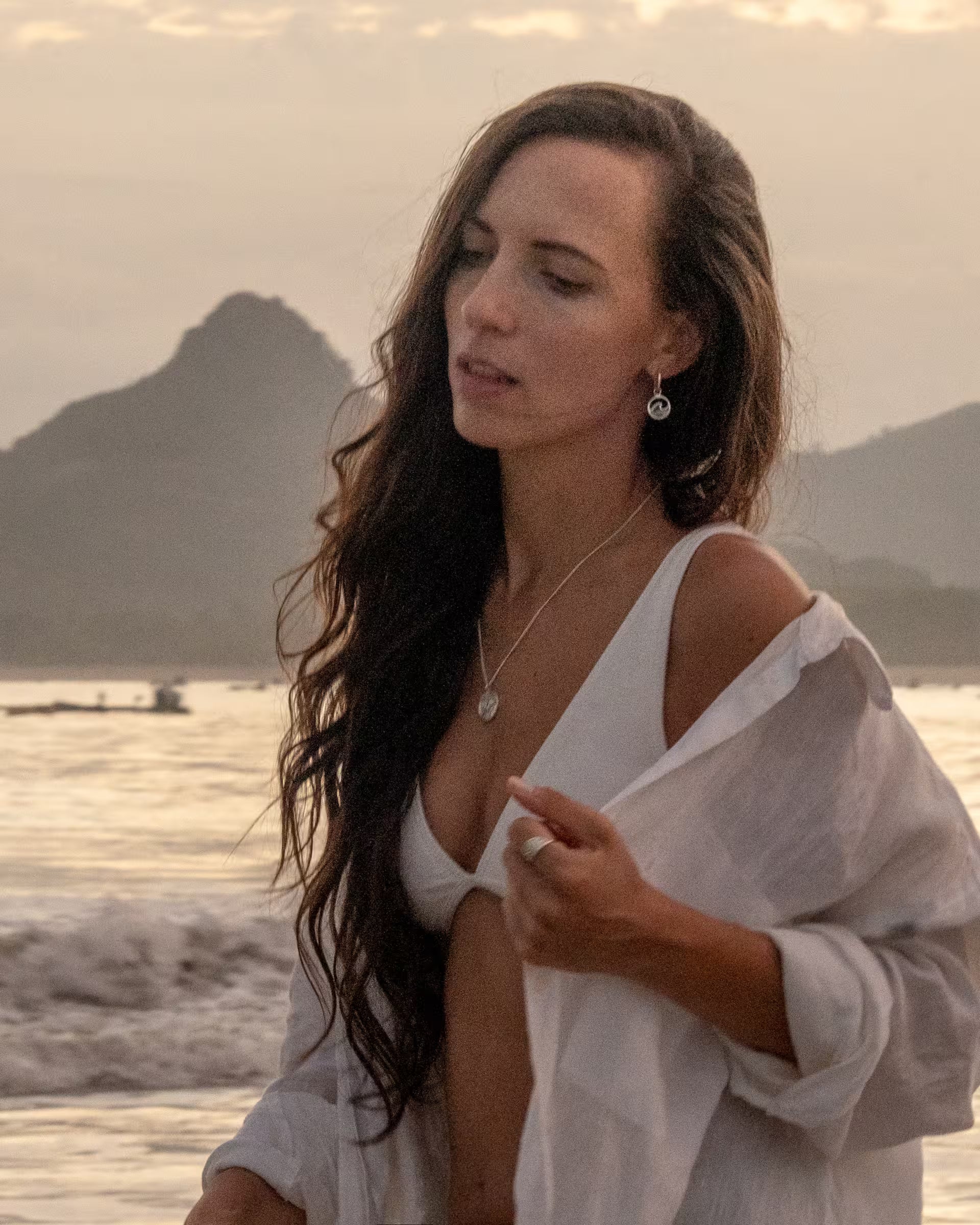 Auf Bali handgefertigte LAJU Creolen Ohrringe mit schimmernder Abalone-Muschel und GILI Paradiese Island Silberkette von der Schmuckmarke ILUH, getragen von einer Frau mit langen, lockigen Haaren im weißen Hemd am Strand bei Sonnenuntergang; die verträumte Atmosphäre und das körnige Bild im Vintage-Look unterstreichen den natürlichen Beach-Stil.