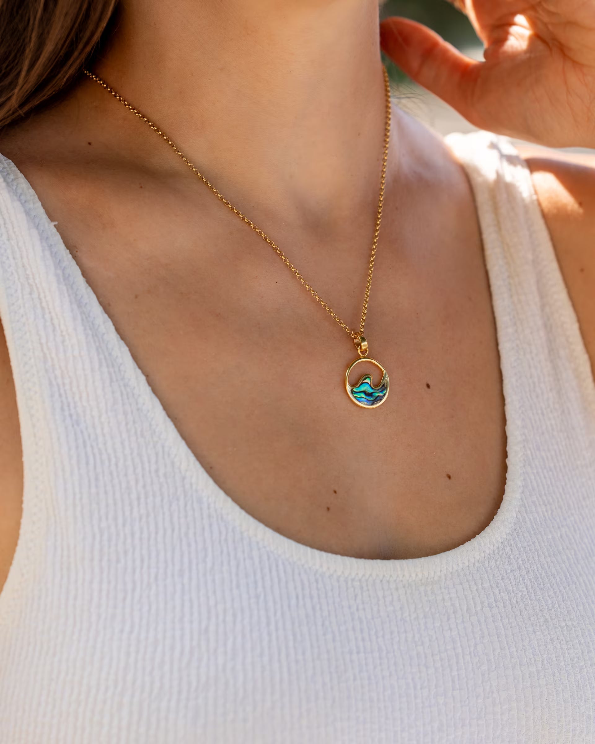 Nahaufnahme im sommerlichen Look von einer Frau im weißen Rippstrick-Oberteil, die die auf Bali handgefertigte goldene LAJU WAVE ABALONE KETTE mit einem runden Wellen-Anhänger, verziert mit schimmernder Abalone-Muschel, aus 18k vergoldetem 925er Silber von der Schmuckmarke ILUH trägt.