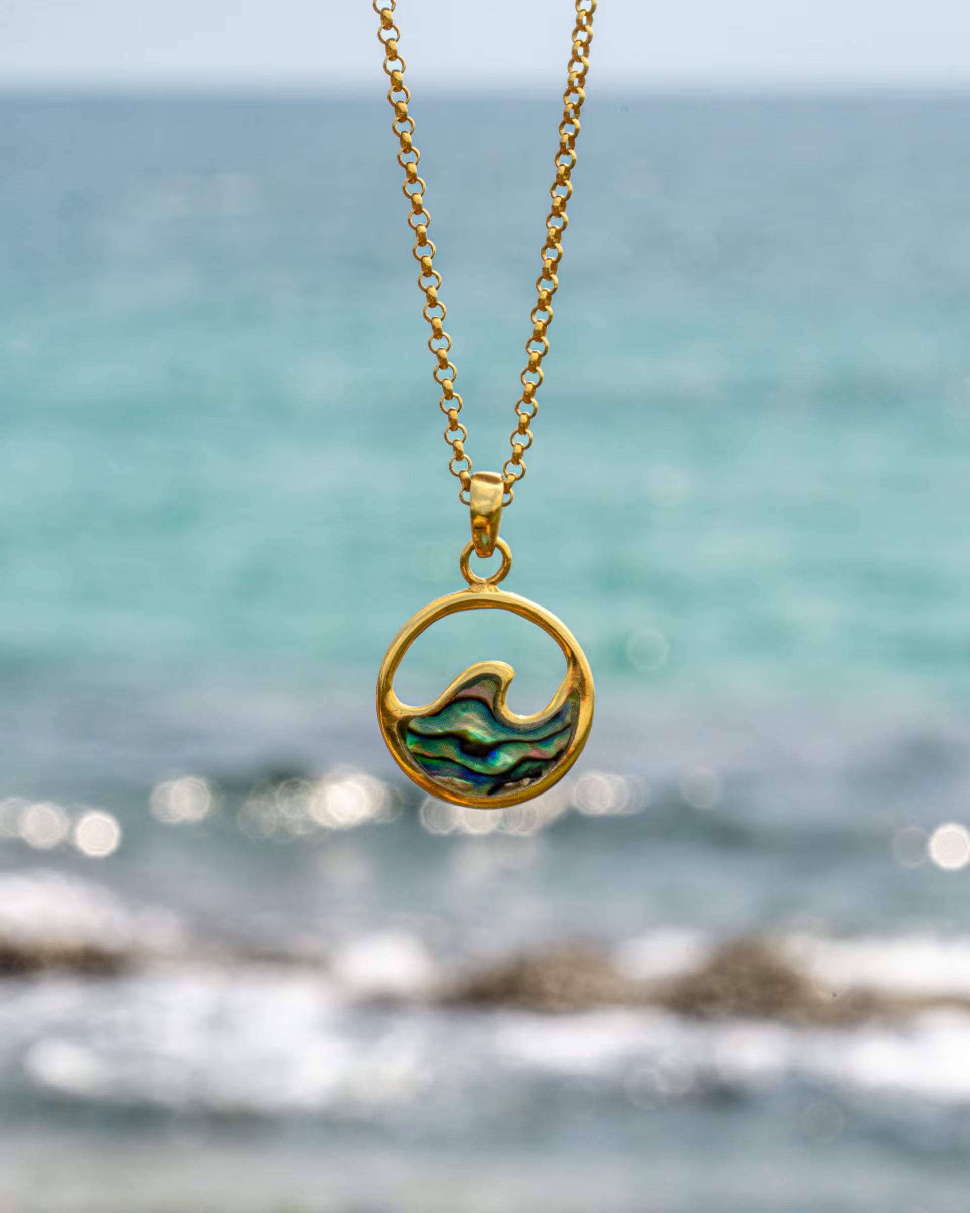 Produktfoto im maritimen Stil von der auf Bali handgefertigten goldenen LAJU WAVE ABALONE KETTE mit einem runden Wellen-Anhänger, verziert mit schimmernder Abalone-Muschel, aus 18k vergoldetem 925er Silber von der Schmuckmarke ILUH, im Hintergrund das leicht unscharfe, türkisblaue Meer mit Schaumkronen.