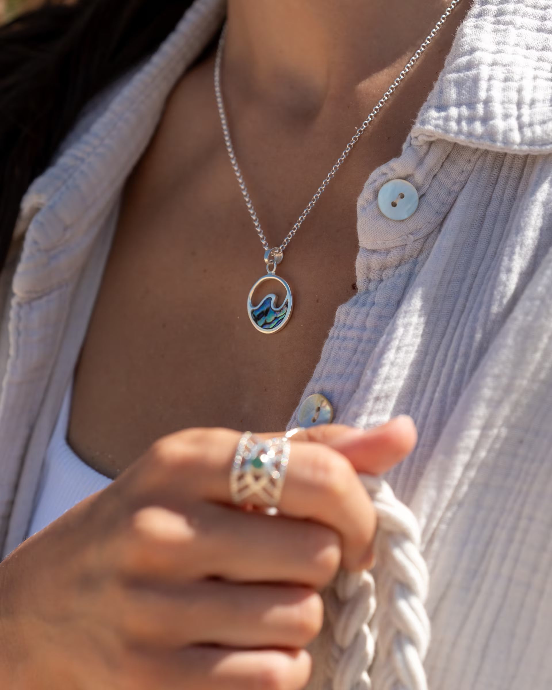 Nahaufnahme im sommerlichen Boho-Look von einer Frau in einer hellen Musselin-Bluse, die die geflochtenen Henkel einer Tasche hält und eine Kombination aus auf Bali handgefertigtem silbernem Schmuck – darunter die LAJU WAVE ABALONE KETTE mit einem runden Wellen-Anhänger aus schimmernder Abalone-Muschel sowie den offenen INTUITION RING mit grünem Smaragd Edelstein – aus 925er Silber von der Schmuckmarke ILUH trägt.