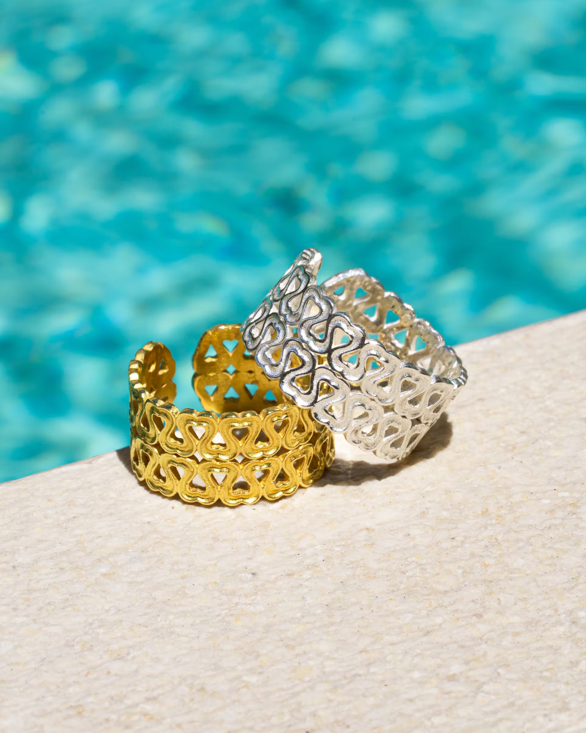 Produktfoto im sommerlichen Stil von zwei auf Bali handgefertigten ENDLESS LOVE Ringen mit kleinen Herz-Muster – einer in Silber aus 925er Silber und einer in Gold aus 18k vergoldetem Silber – von der Schmuckmarke ILUH, platziert am Poolrand vor türkisfarbenem Wasser.