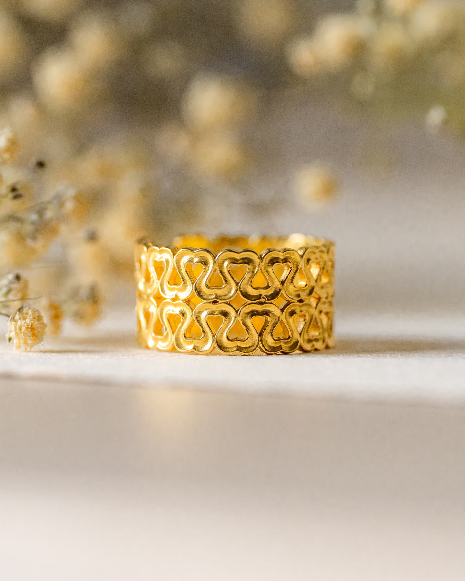Auf Bali handgefertigter goldener ENDLESS LOVE HEARTS Ring mit Herz-Muster aus 18k vergoldetem 925er Silber von der Schmuckmarke ILUH, als Produktfoto vor einem hellen Hintergrund mit verschwommenen Trockenblumen.