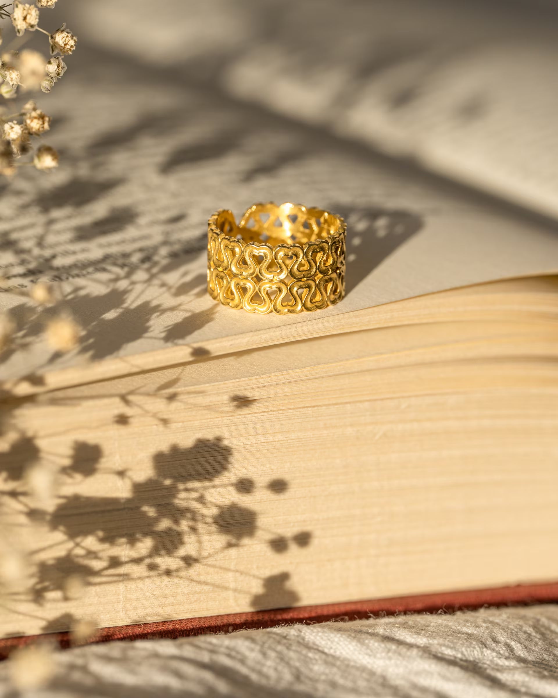 Produktfoto im romantischen Stil von dem auf Bali handgefertigten goldenen ENDLESS LOVE RING mit ausgestanztem Herz-Muster aus 18k vergoldetem 925er Silber von der Schmuckmarke ILUH, elegant platziert auf einem offenen Buch im warmen Sonnenlicht mit floralen Schatten von Trockenblumen.